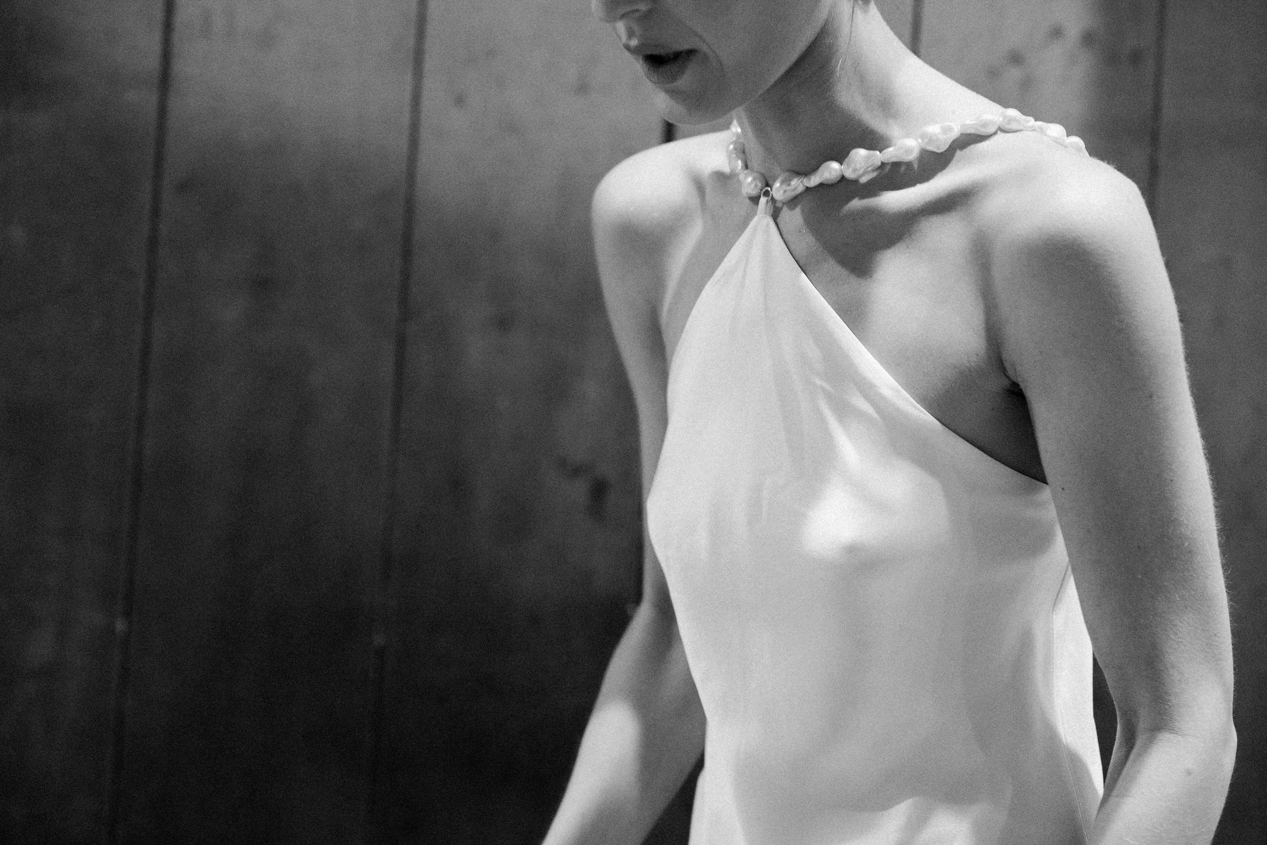 Dettaglio di una donna in abito bianco con collana di perle, vista parziale, in bianco e nero.