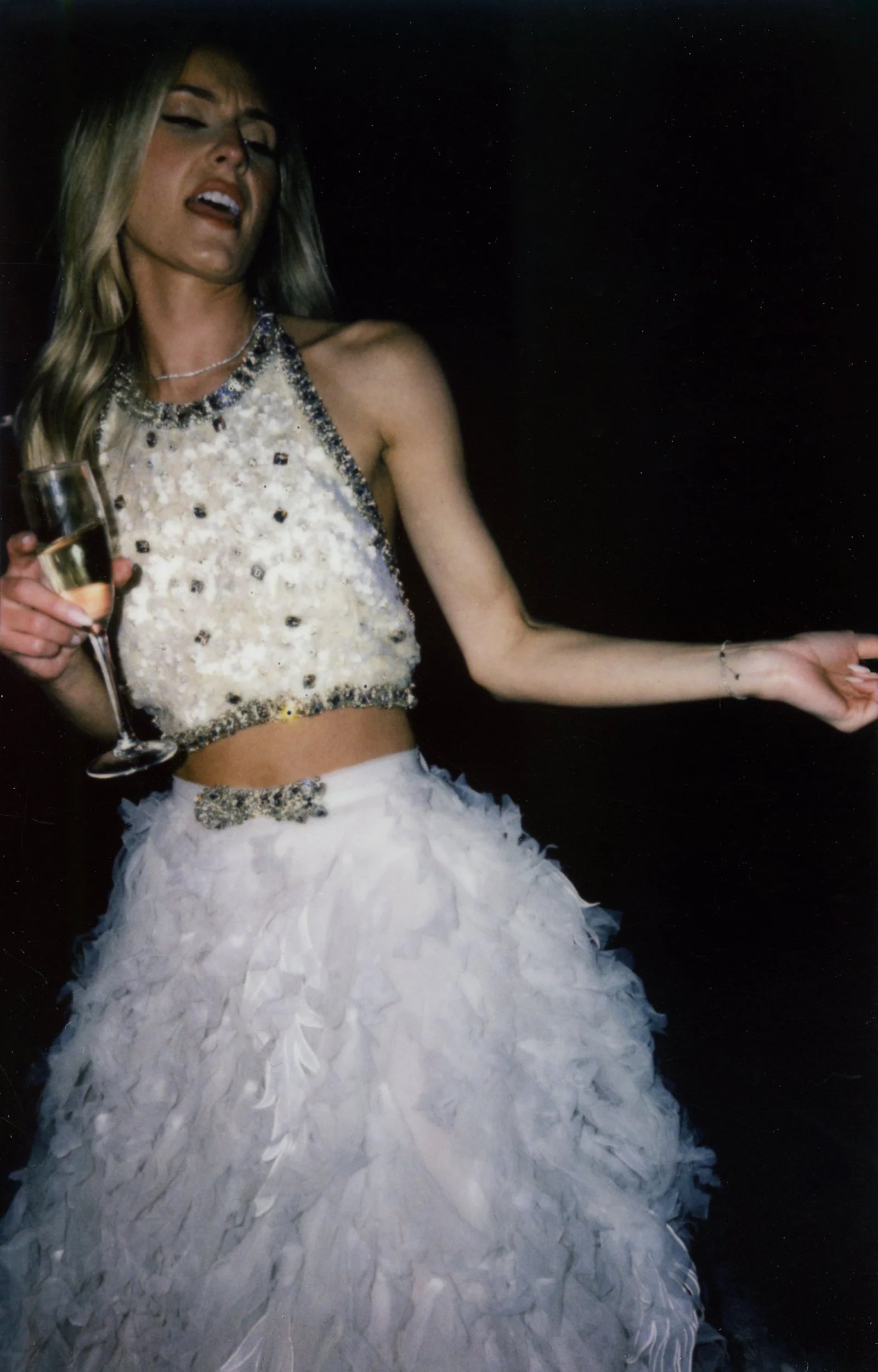 Donna con abito da sera bianco decorato con paillettes e dettagli in cristallo, che tiene un bicchiere di champagne in una mano, in un ambiente scuro.
