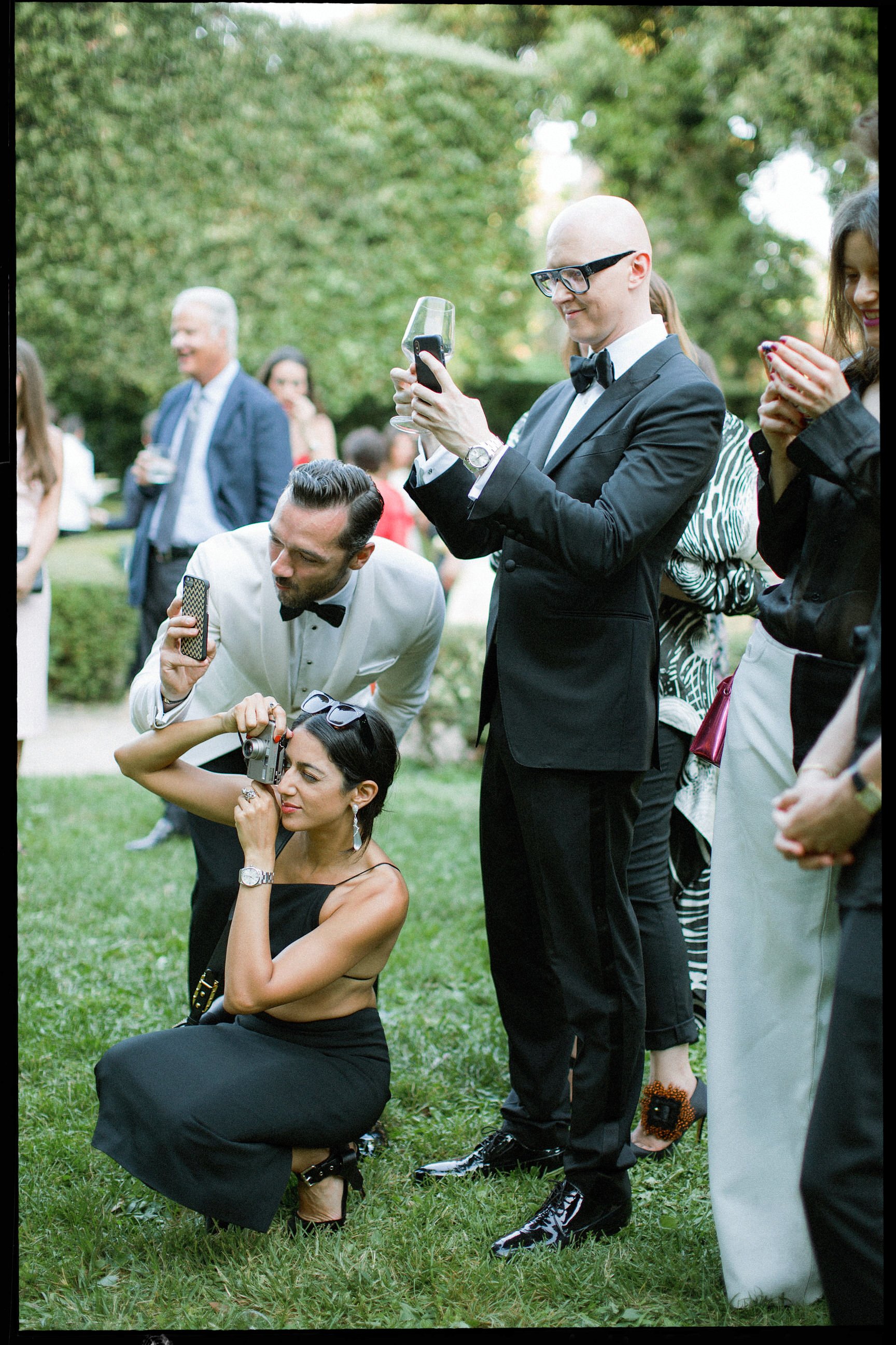 Giornata sociale in giardino con persone formali, alcune che scattano foto, tra cui un uomo in smoking con cappotto nero, una donna in abito nero seduta a terra, altre persone che chiacchierano e si tengono impegnate, alberi verdi sullo sfondo.