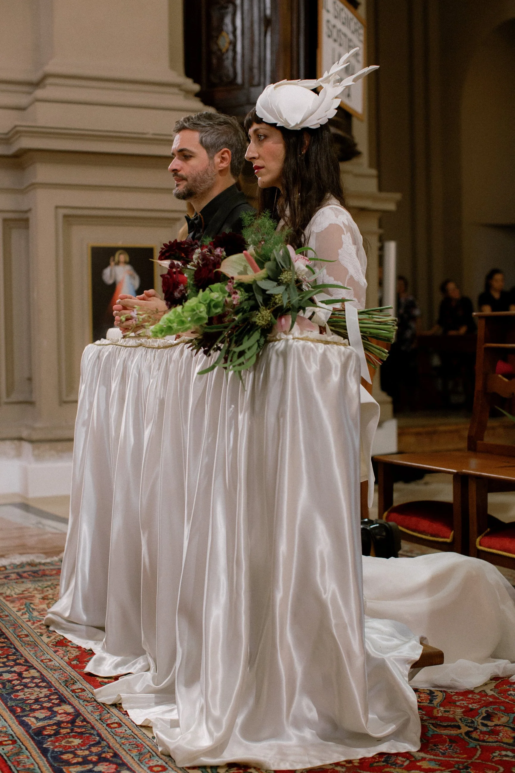 Un uomo e una donna stanno partecipando a un matrimonio religioso. La donna indossa un abito bianco con un cappellino decorato con piume e tiene un bouquet di fiori. L'uomo indossa un abito scuro e una cravatta nera.