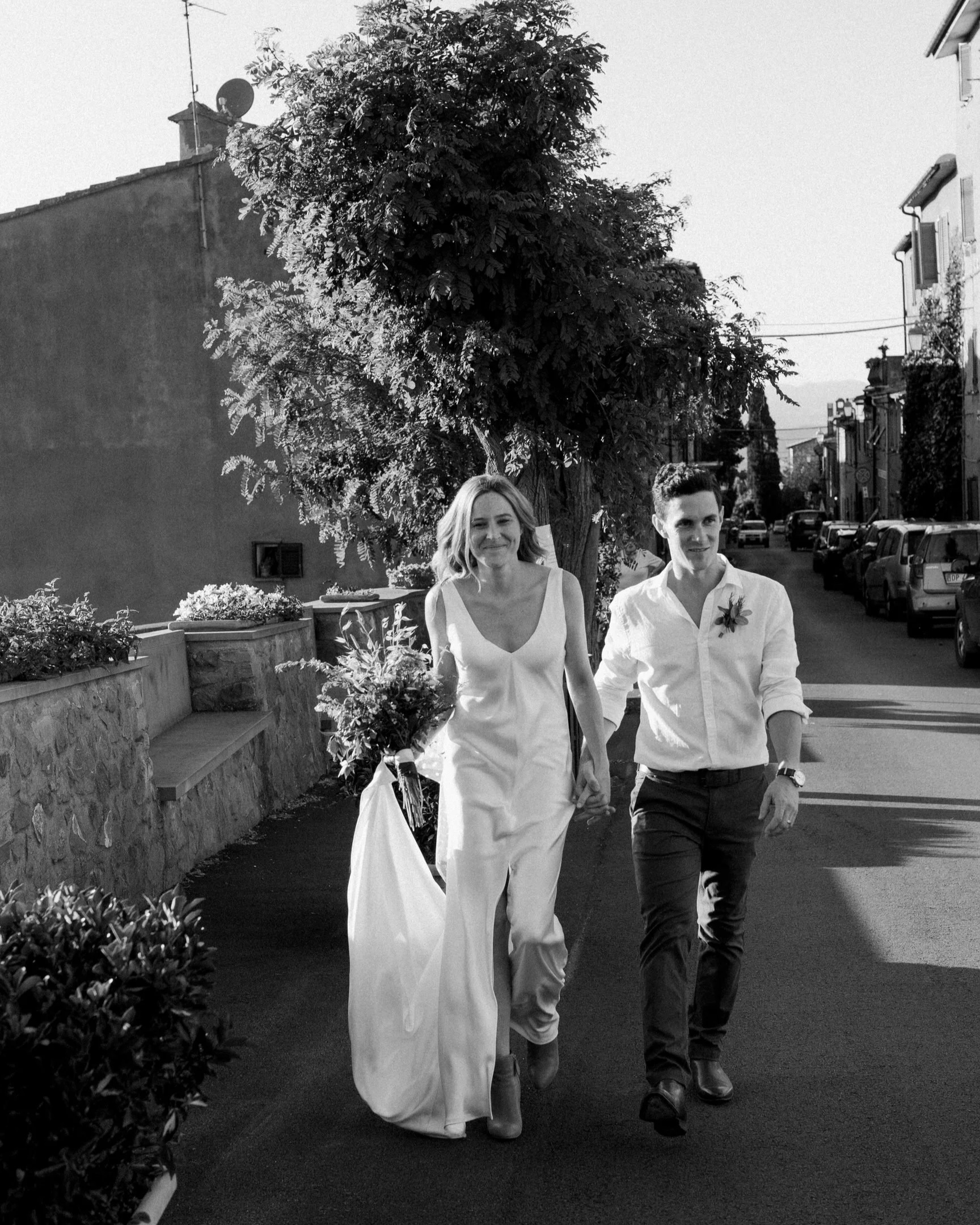 Una donna e un uomo che camminano mano nella mano lungo una strada di città. La donna indossa un abito bianco e tiene un bouquet di fiori, probabilmente in occasione di un matrimonio, mentre l'uomo è vestito con una camicia bianca e pantaloni scuri.
