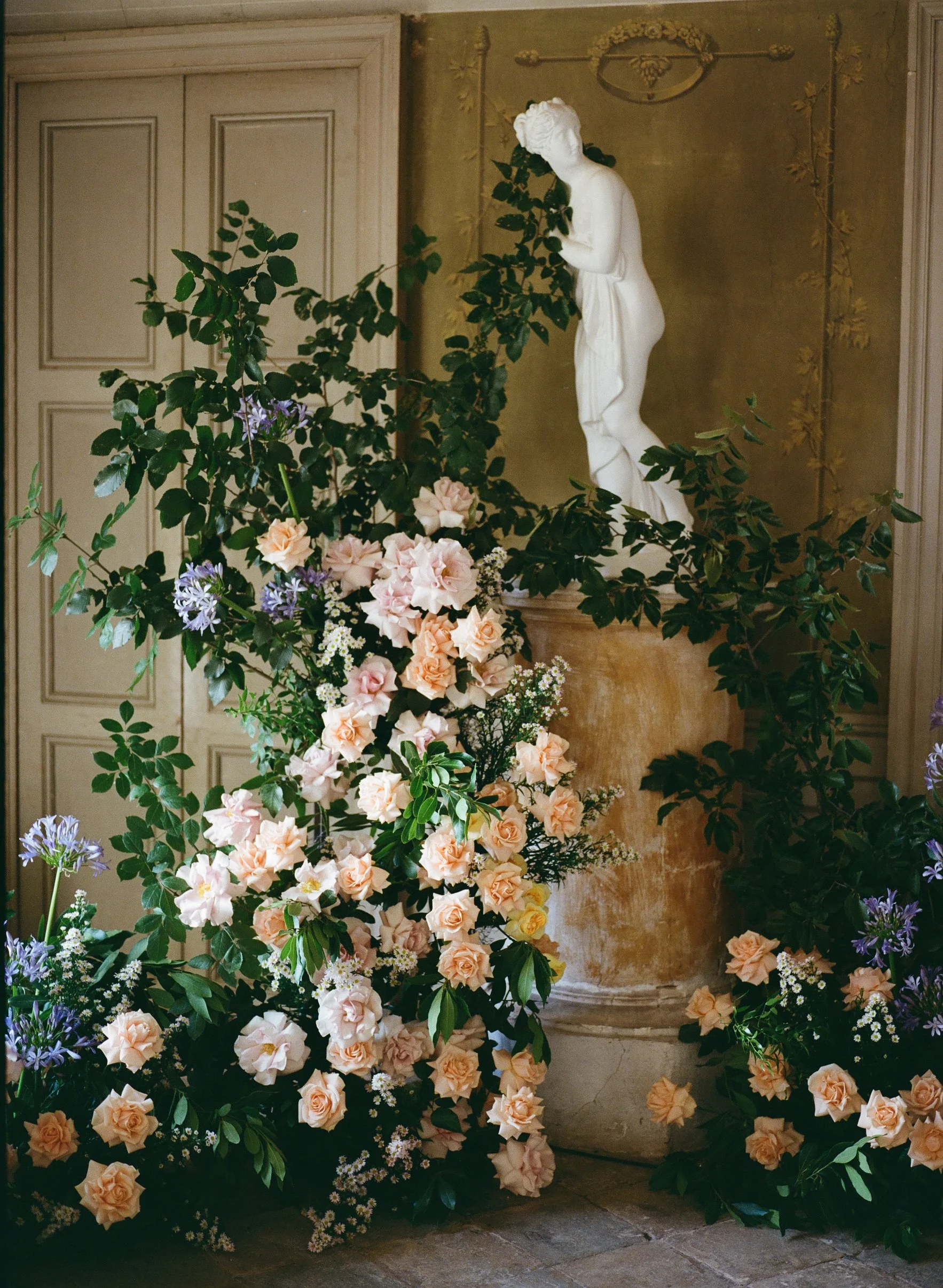 Ornamento di fiori e piante color pastello davanti a una statua di scultura in stile classico, posizionata in un ambiente con decorazioni classiche.