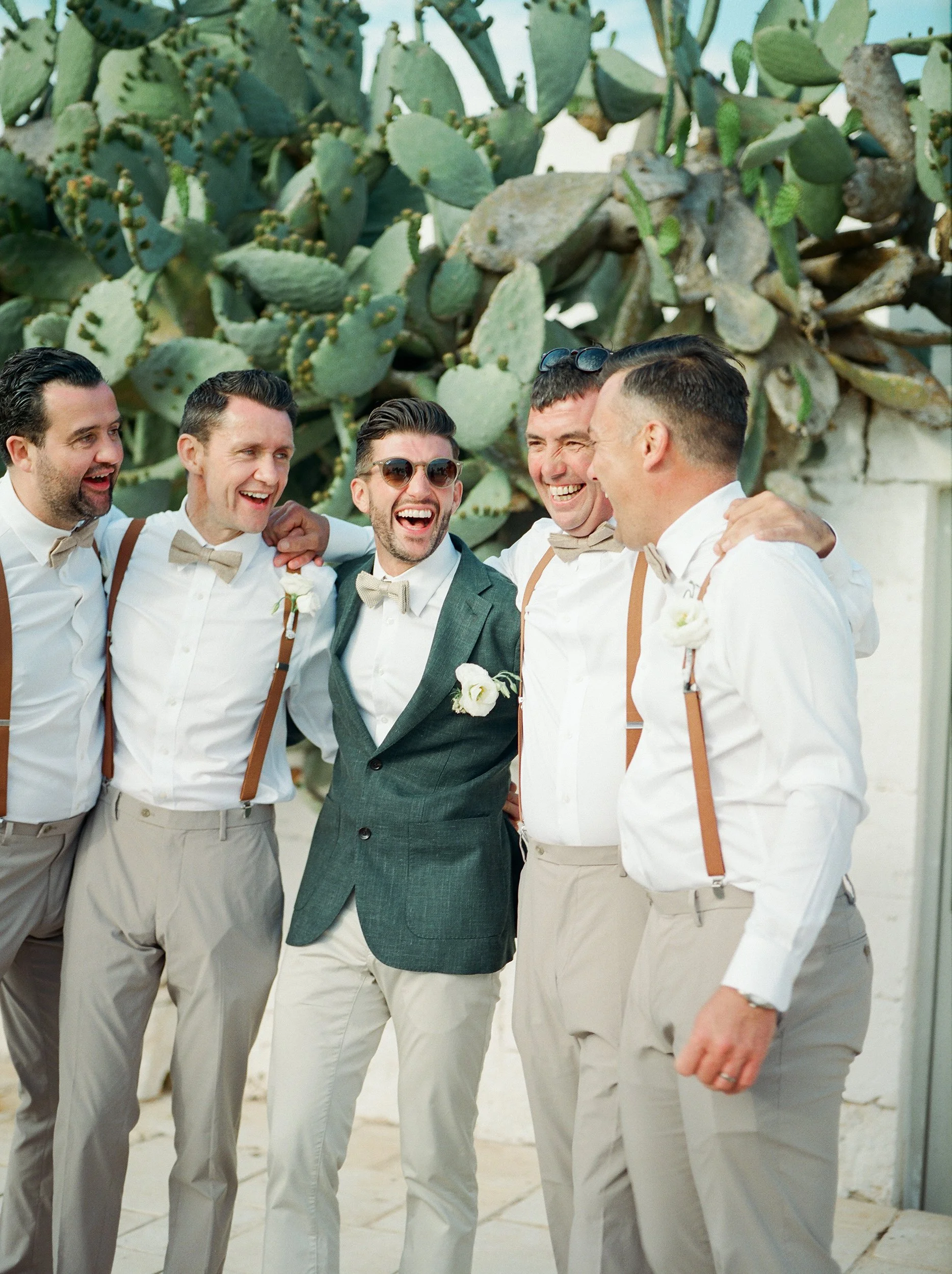 Un gruppo di uomini vestiti elegantemente, sorridenti e che si abbracciano, si trovano all'aperto con un grande cactus sullo sfondo.