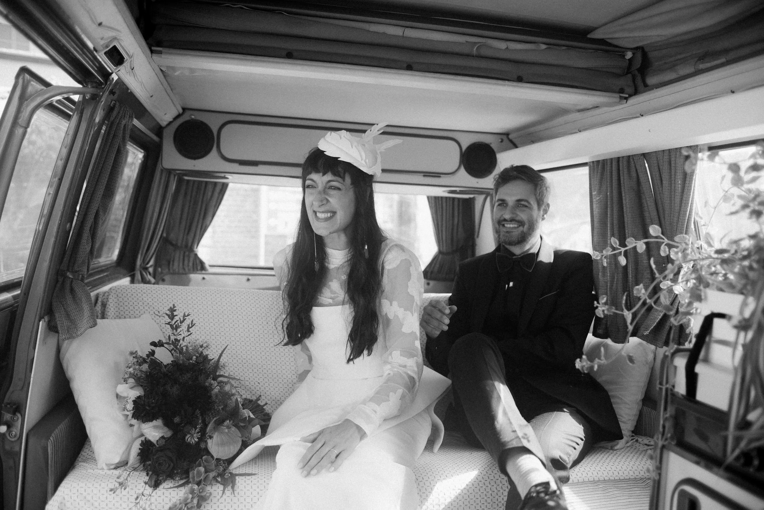 Coppia sorridente su un letto in un camper, con lei che indossa un cappello di carta e tiene un mazzo di fiori, lui vestito elegante con abito e farfalla, caoticamente seduti con tende alle finestre e decorazioni