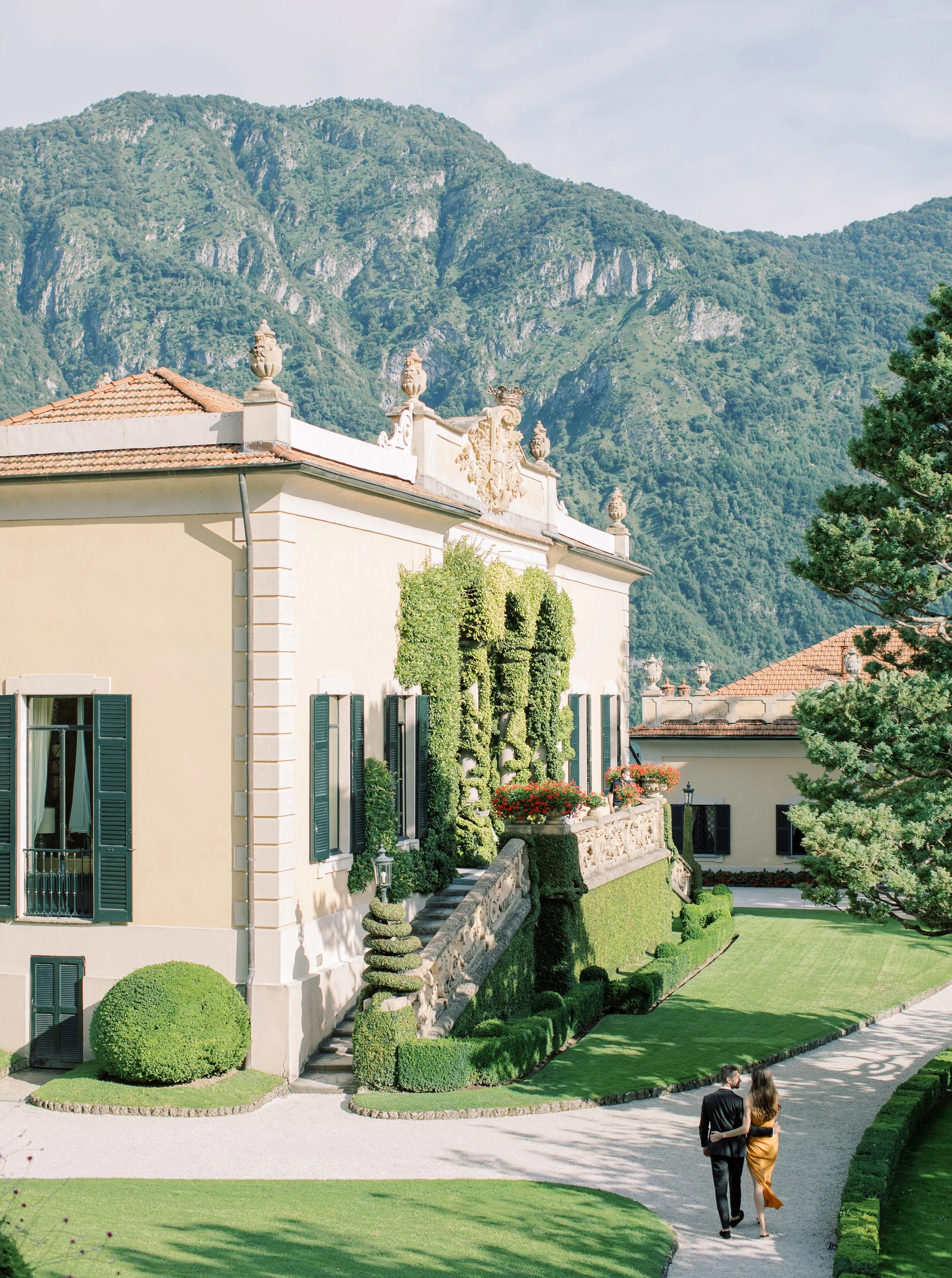 Giardino ben curato con prato verde, siepi e piante decorative, davanti a un edificio storico con decorazioni e sculture, con montagne sullo sfondo. Due persone vestite eleganti camminano lungo il percorso.