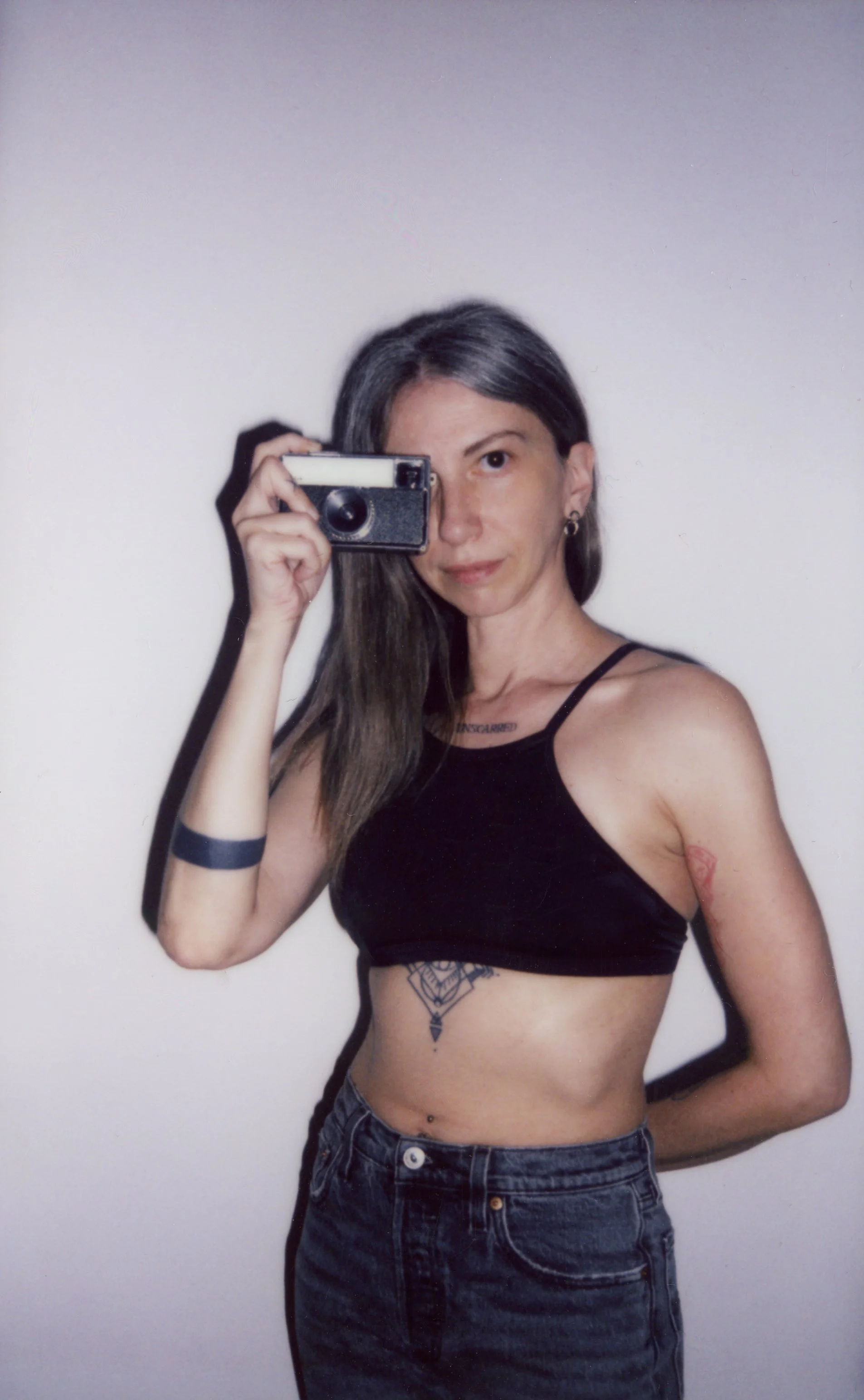 Una donna con capelli castani lunghi, occhi castani e pelle chiara che indossa una maglietta senza maniche nera e jeans, si sta prendendo una selfie di fronte a uno sfondo neutro con una macchina fotografica economica. Ha vari tatuaggi visibili su braccia, addome e torace.