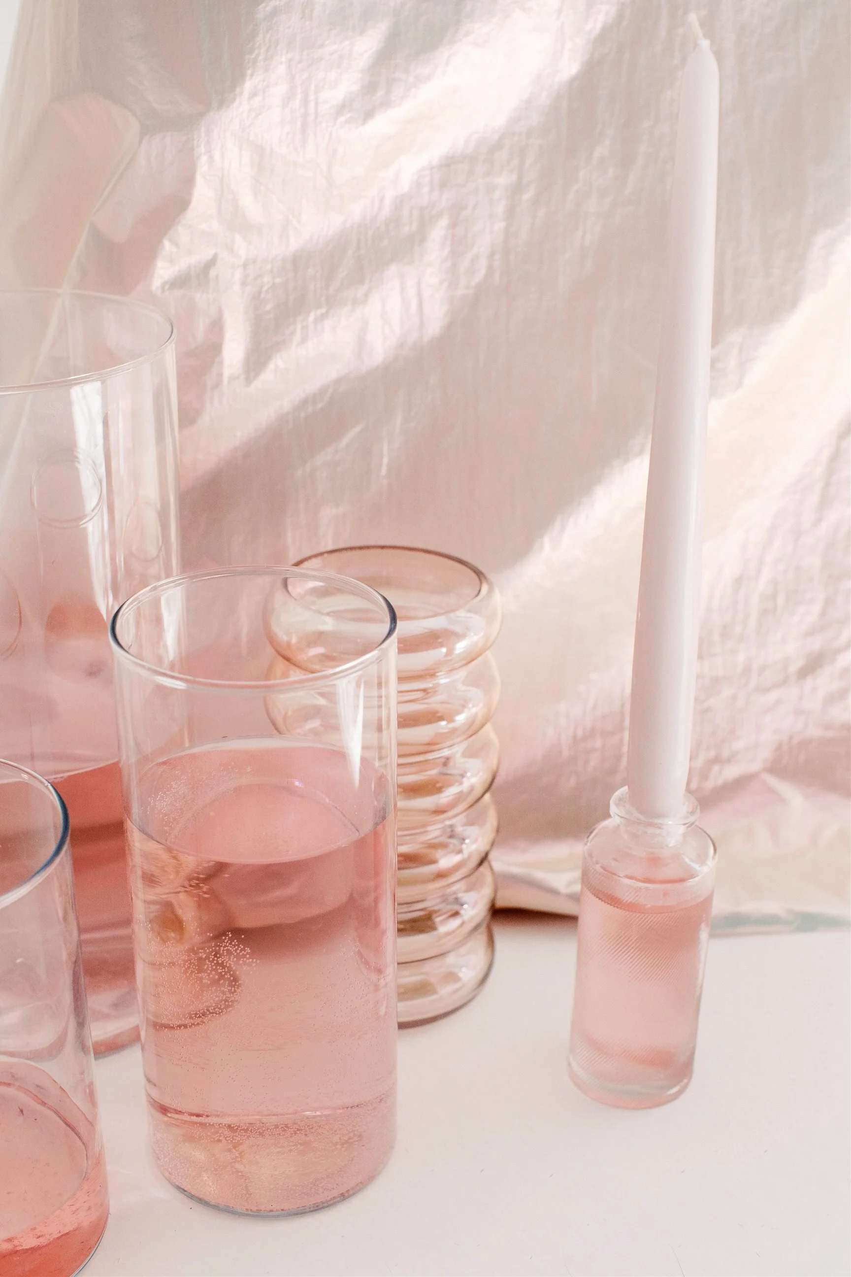 Vasi di vetro riempiti con una sostanza rosa trasparente, un candeliere bianco con candela bianca, sfondo di tessuto bianco con luci soffuse.