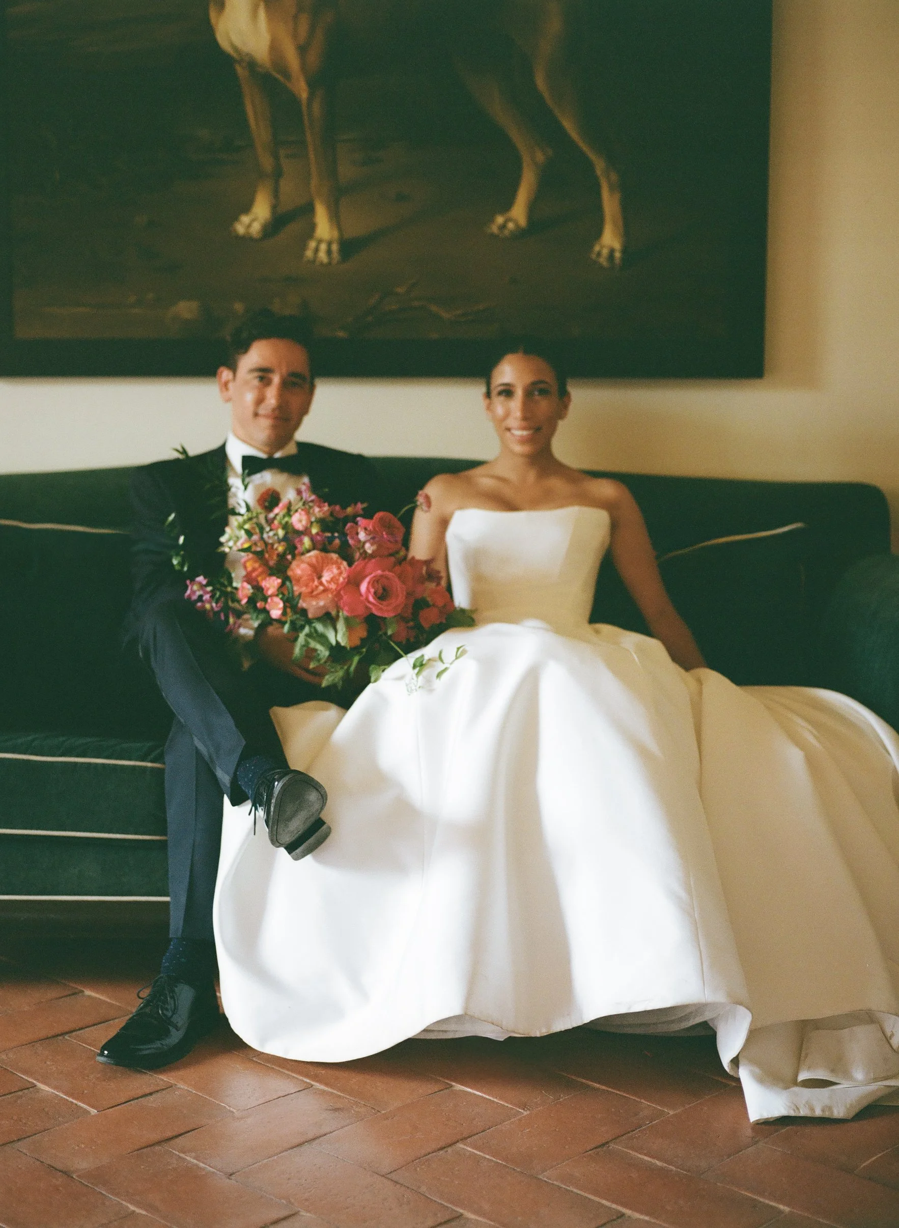 Una coppia di sposi seduta su un divano con la sposa in abito bianco e il marito in smoking nero, entrambi sorridenti, con un mazzo di fiori rosa e rossi in mano.
