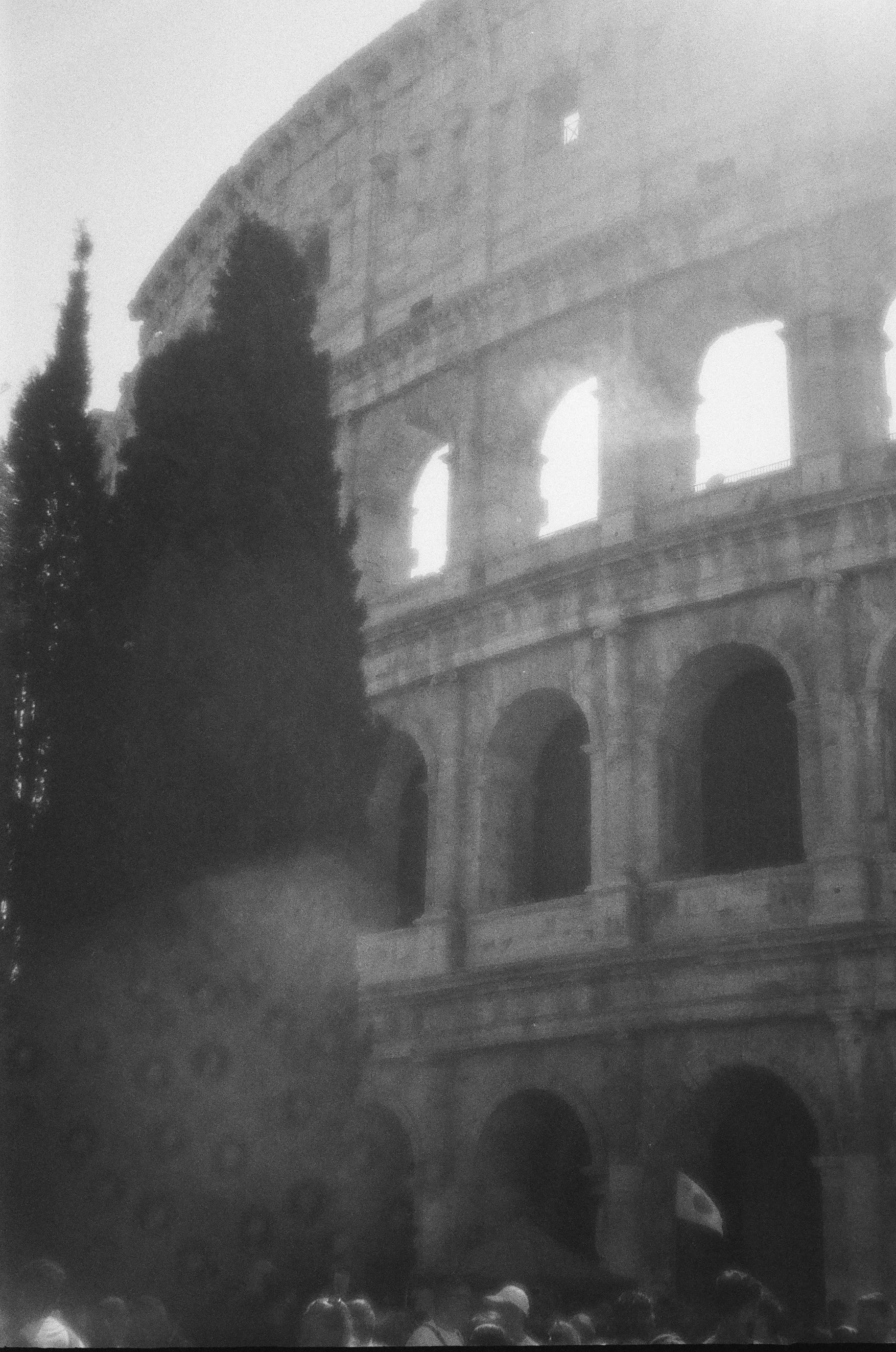 Veduta parziale del Colosseo di Roma visto attraverso una fitta nebbia, con alcune sagome di persone in primo piano.