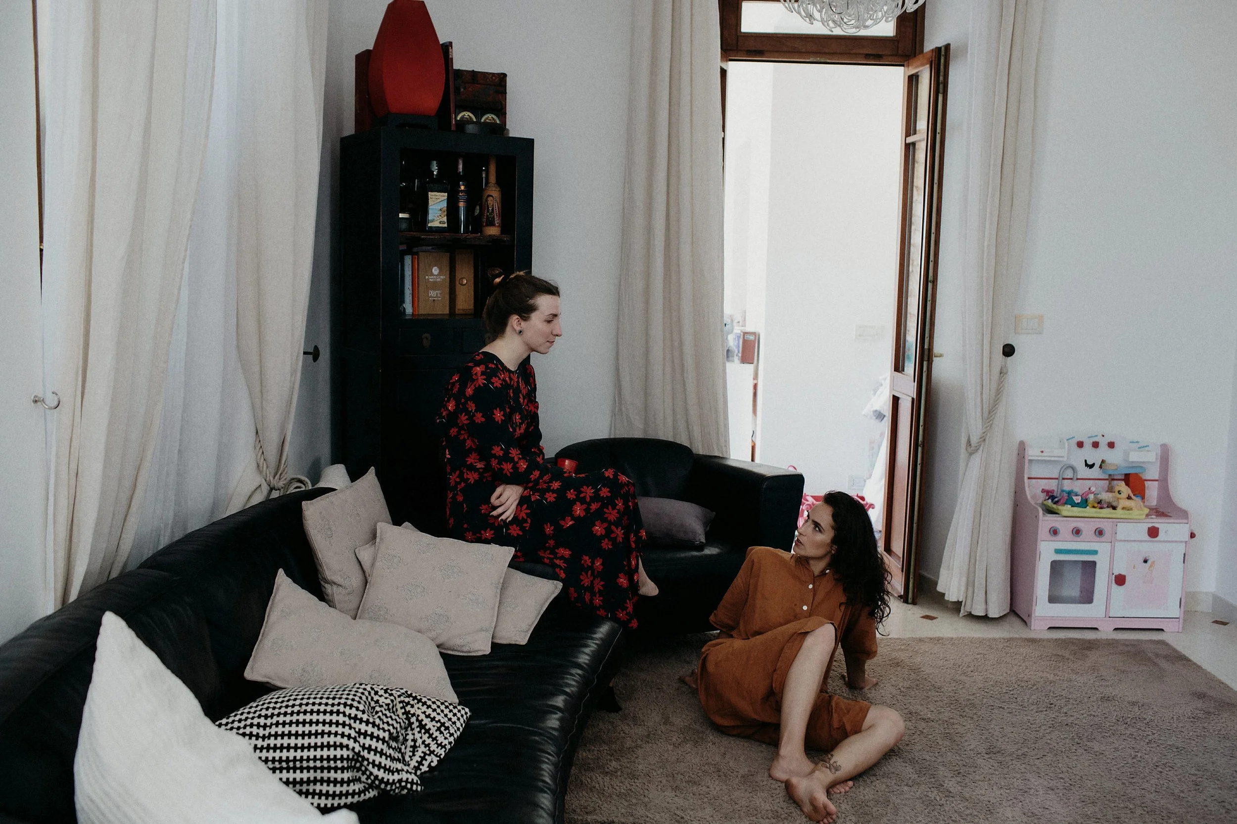 Tre donne sedute e a terra in un soggiorno, una sulla poltrona, una sulla poltrona e una sul pavimento, parlano tra loro. La stanza ha tende chiare, una libreria con oggetti e libri, e una cucina in parte visibile.