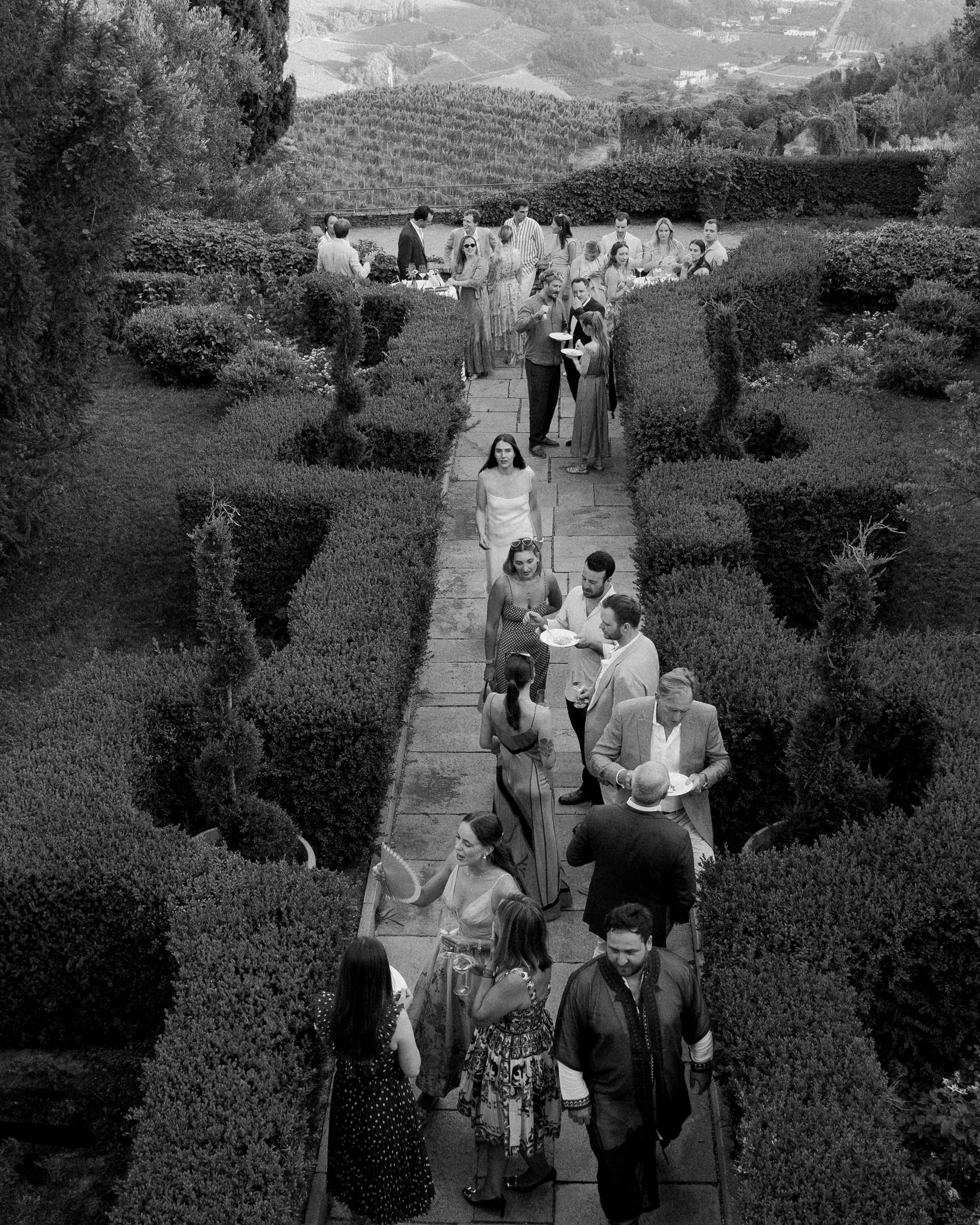Un gruppo di persone in un'elegante riunione in un giardino con vista collinare, molte con calici di vino, alcune che parlano, altre che si muovono lungo un vialetto.