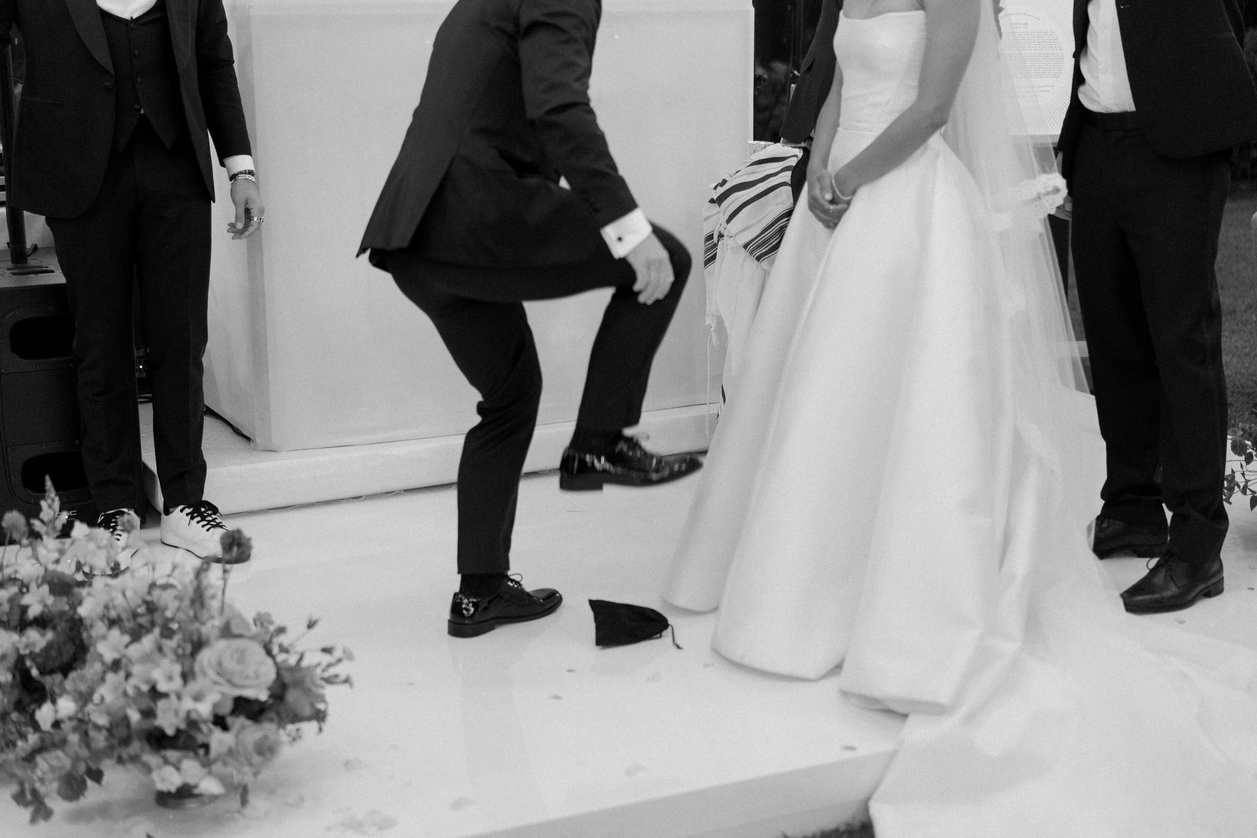 Abito tradizionale da sposa bianco con gonna ampia, indossato da una donna, in un contesto di matrimonio.

Uomini in abiti formali con giacca e pantaloni scuri, alcune scarpe vistose.

Fronte a una parete bianca, con dettagli di decorazioni floreali 