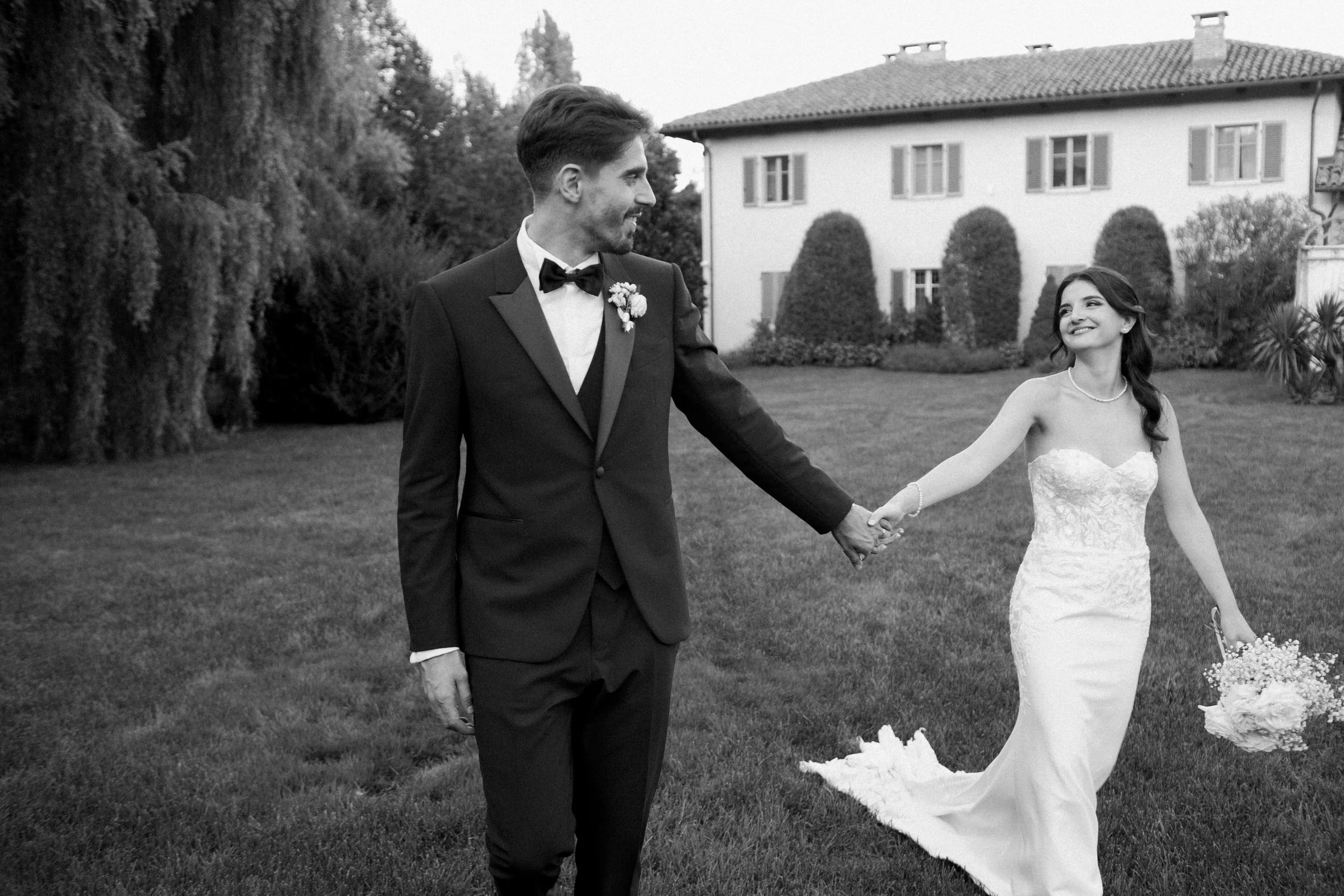 Due persone che si tengono per mano, un uomo in smoking e una donna in abito da sposa, camminano in un giardino di fronte a una casa grande con finestre e cespugli curati, in un'immagine in bianco e nero.