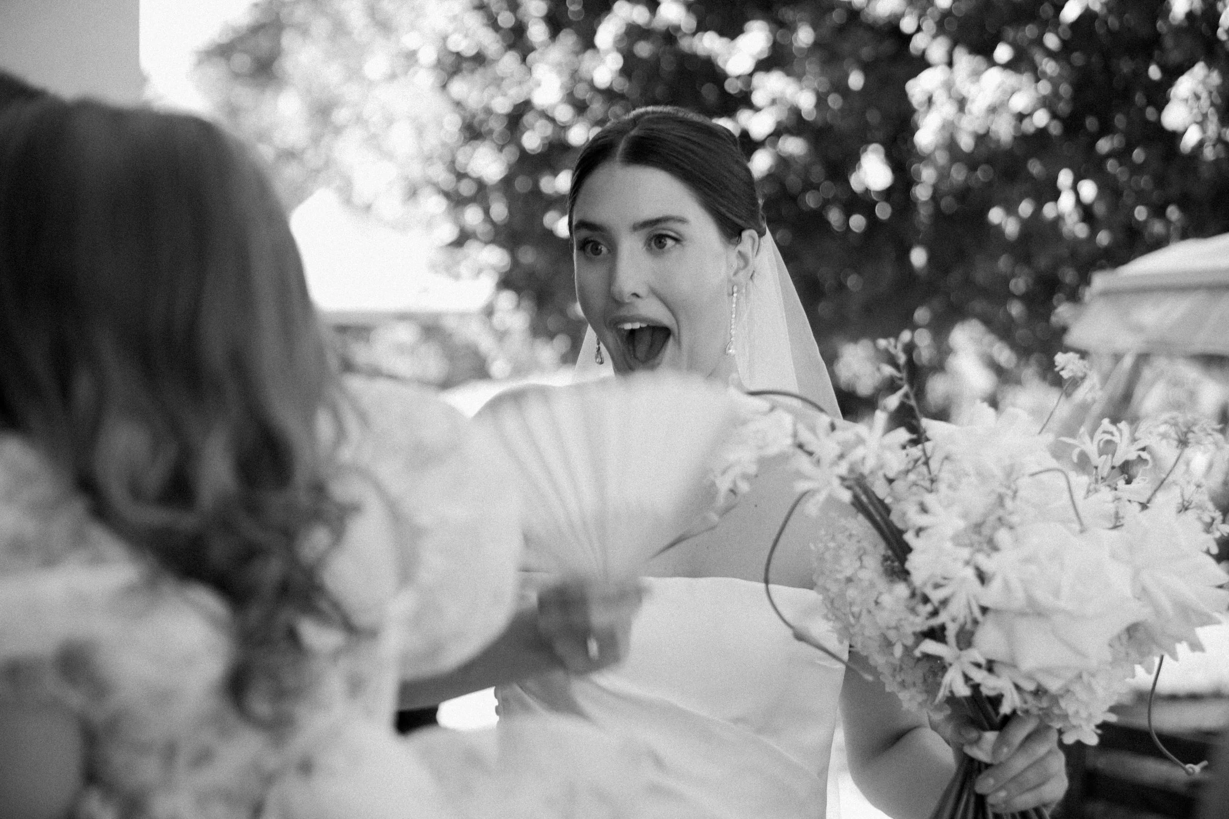 Sposa sorridente con orecchini pendenti, vestita bianca, che tiene un bouquet di fiori durante un matrimonio all'aperto, con sfondo di alberi e luce naturale.