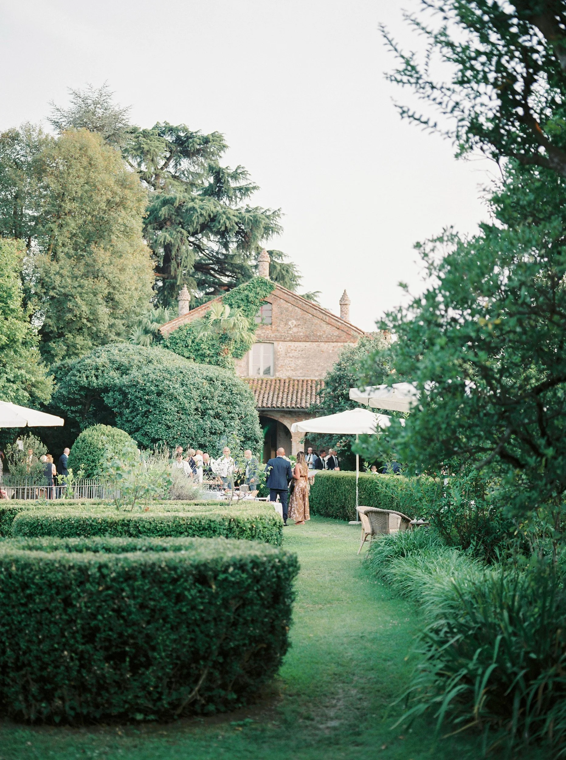 Una riunione elegante si svolge in un giardino decorato con cespugli pieni e ombrelloni, con un'antica casa in pietra sullo sfondo, circondata da alberi.