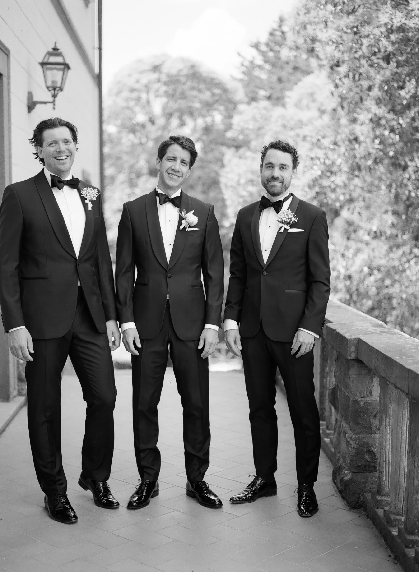 Quattro uomini vestiti in abiti da cerimonia, sorridenti, che camminano all'aperto, con uno sfondo di alberi e un muro in pietra. Sono vestiti con smoking neri, camicie bianche e papillon, decorati con fiori.