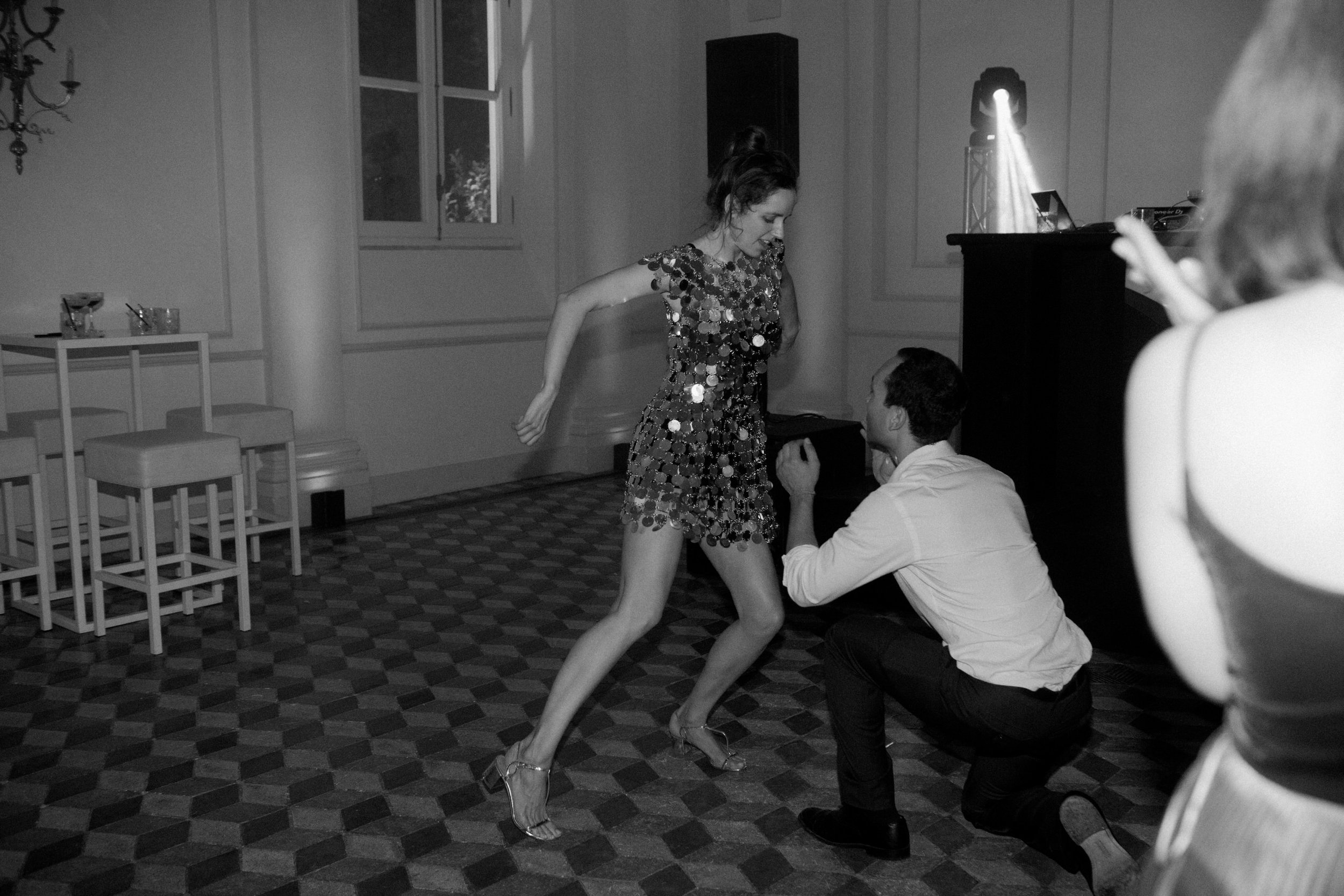 Una donna con un vestito ricoperto di bottoni luccicanti danza con un uomo chino che tiene il suo piede. La scena si svolge in una stanza con tavoli e sedie, con luci del DJ e una finestra in background.