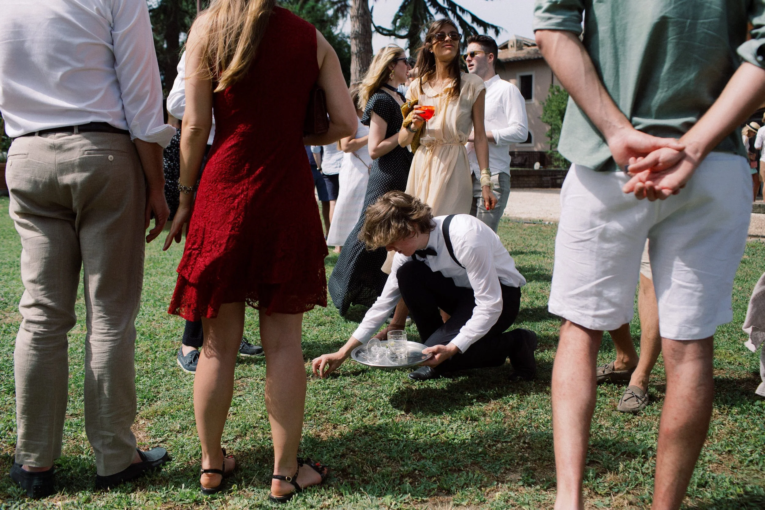 Gruppo di persone in abiti eleganti che partecipano a un evento all'aperto, con alcuni che si chinano e un cameriere vestito di nero con camicia bianca che serve bevande su un vassoio argentato.