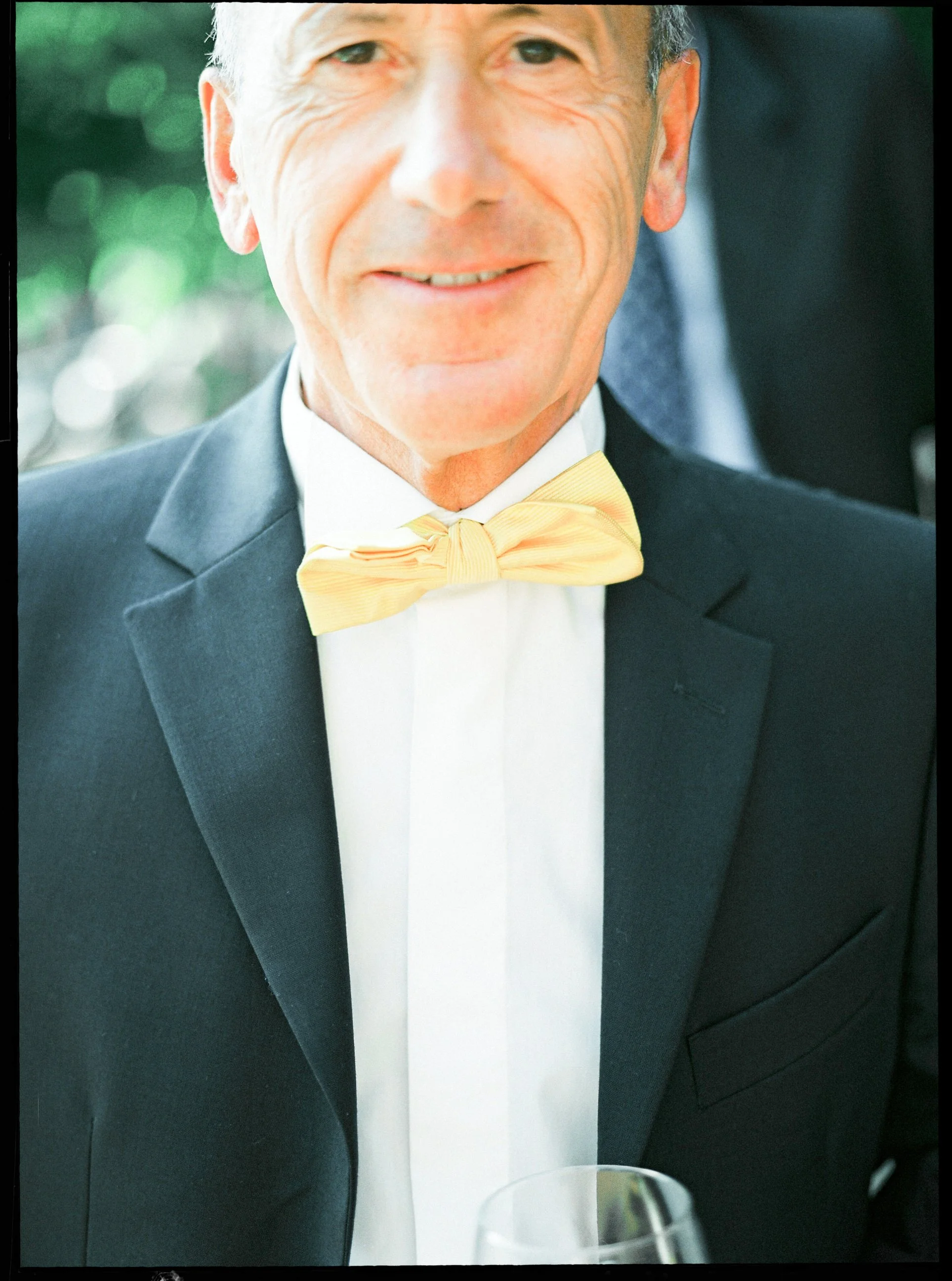 Primo piano di un uomo anziano con capelli grigi, sorriso sottile, indossa un abito nero con camicia bianca e una cravatta gialla, in un contesto elegante.