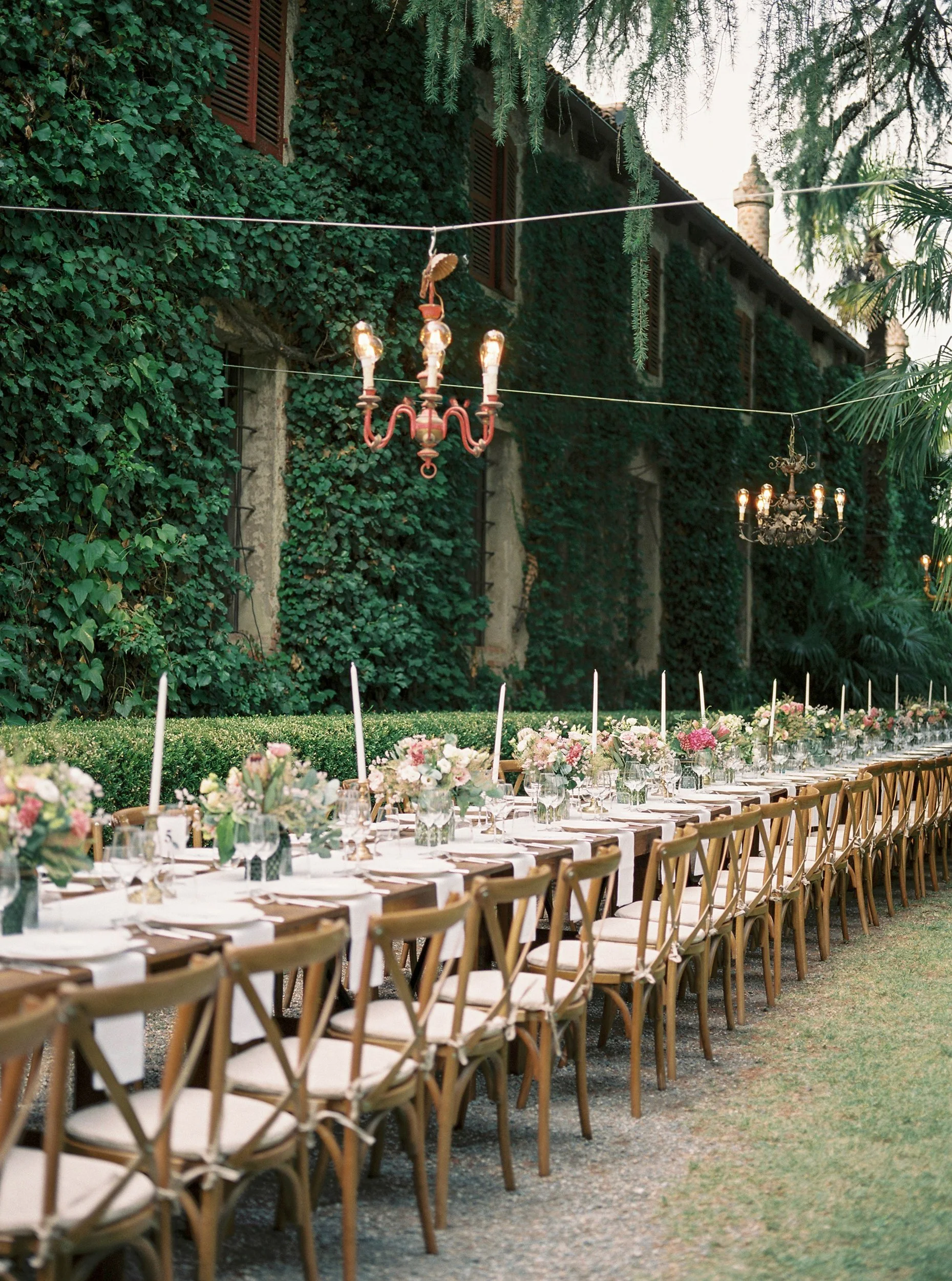 Una lunga tavola decorata con fiori e candele in un giardino esterno, con una parete coperta di edera e un edificio in stile rustico sullo sfondo.