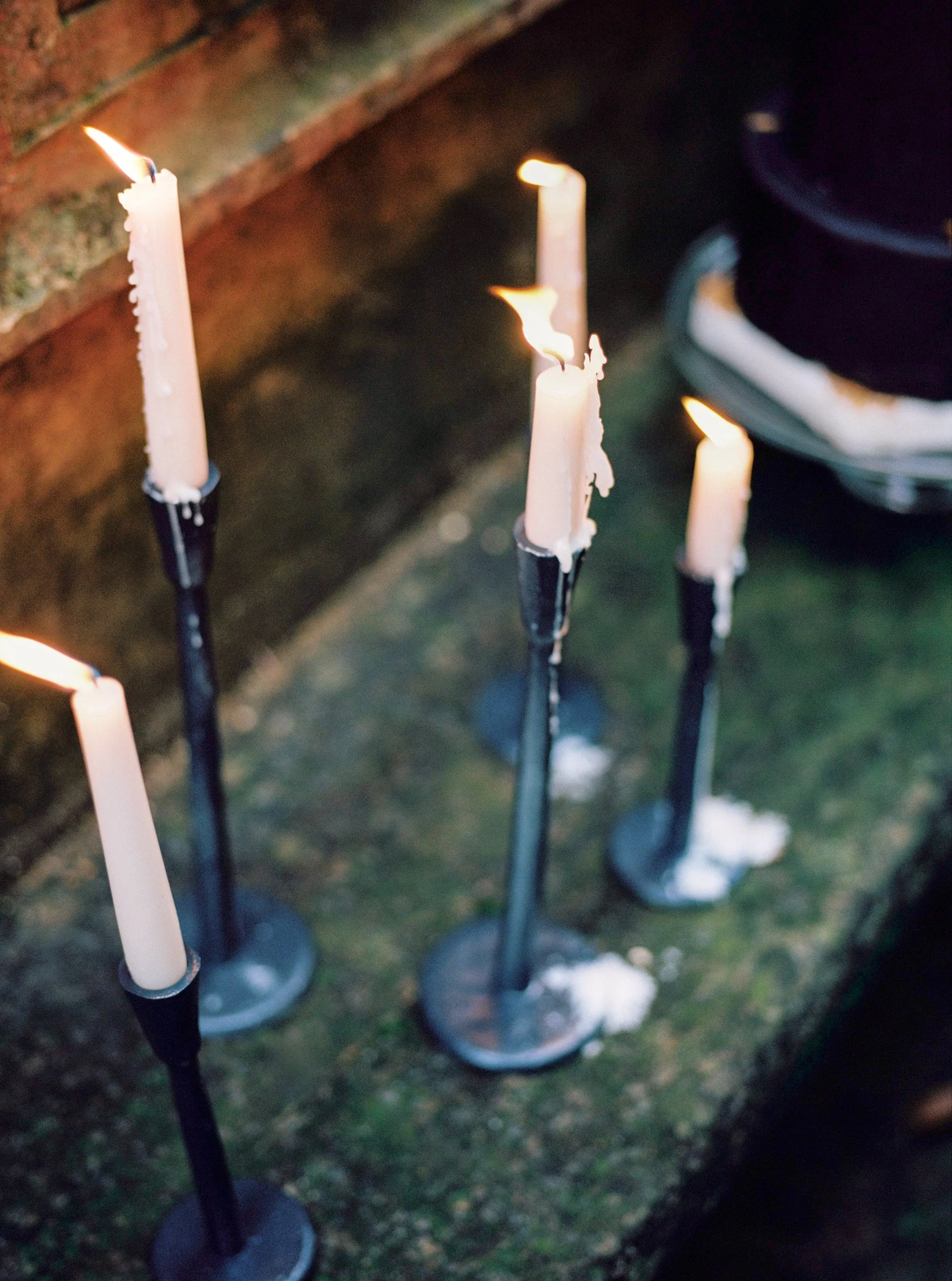 Lucine accese in setting outdoor, disposte su supporti metallici, con cera sciolta e fiamme accese, in un ambiente notturno o correttamente illuminato al buio.