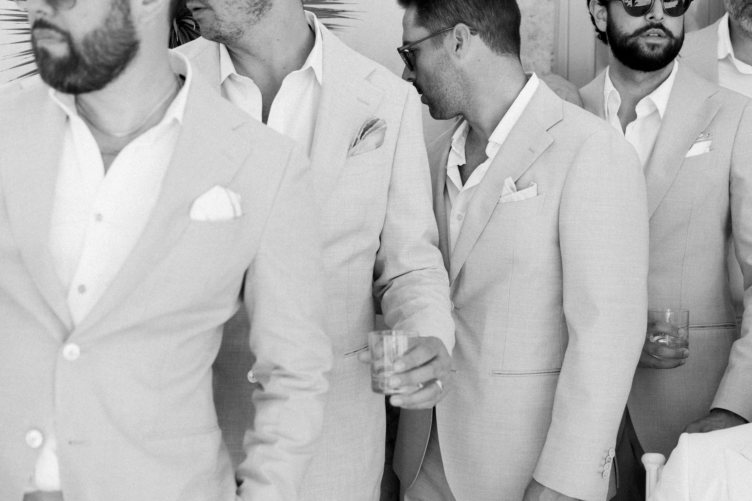 Gruppo di uomini con giacche chiare, alcuni con occhiali scuri, che tengono bicchieri di bevande durante un evento sociale.