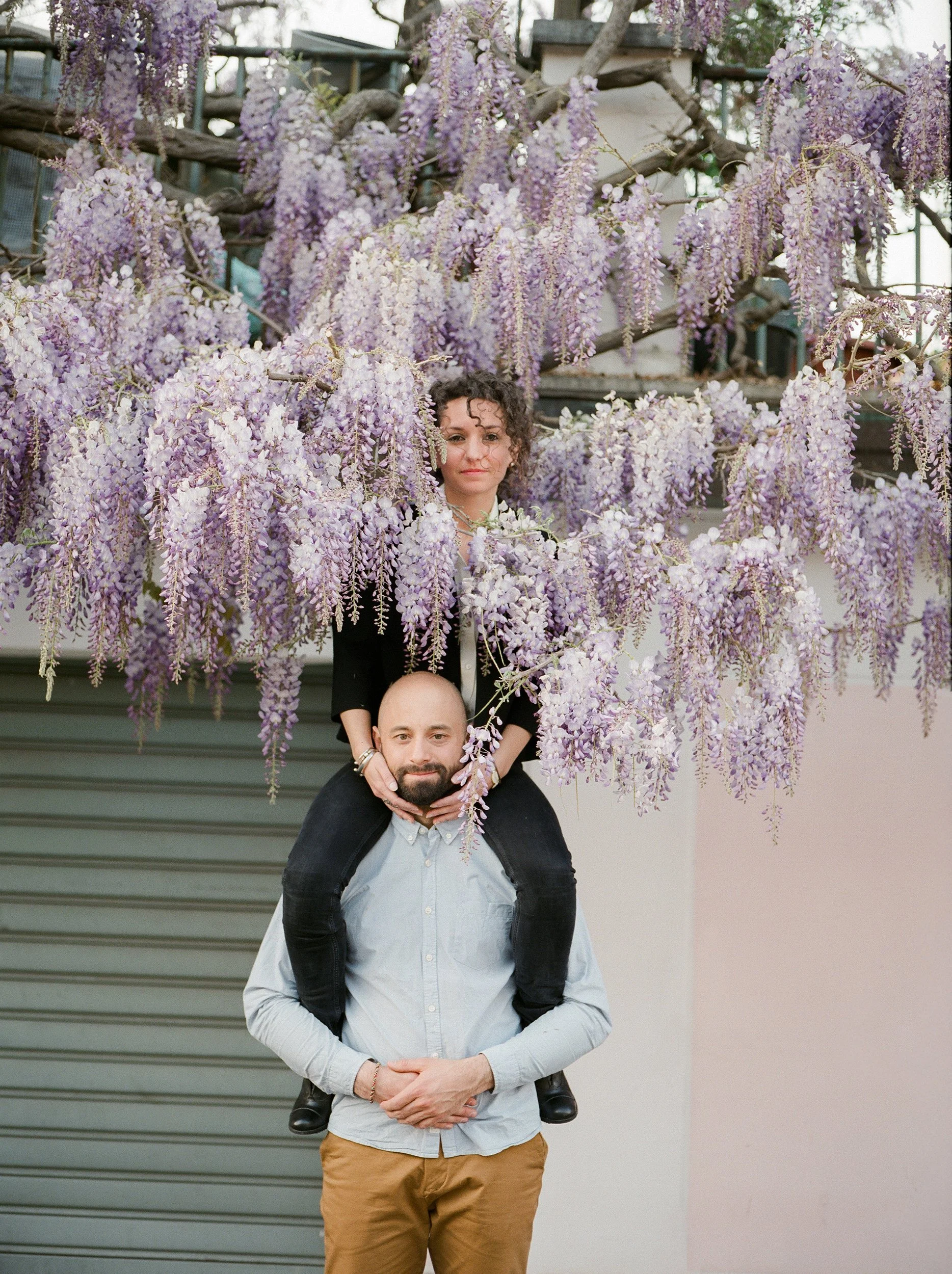 Un uomo con un vestito grigio e pantaloni marroni sta con una donna sulle sue spalle, tra fiori di glicine viola e bianco. Sullo sfondo, una porta garage di colore grigio ed un muro di colore chiaro.
