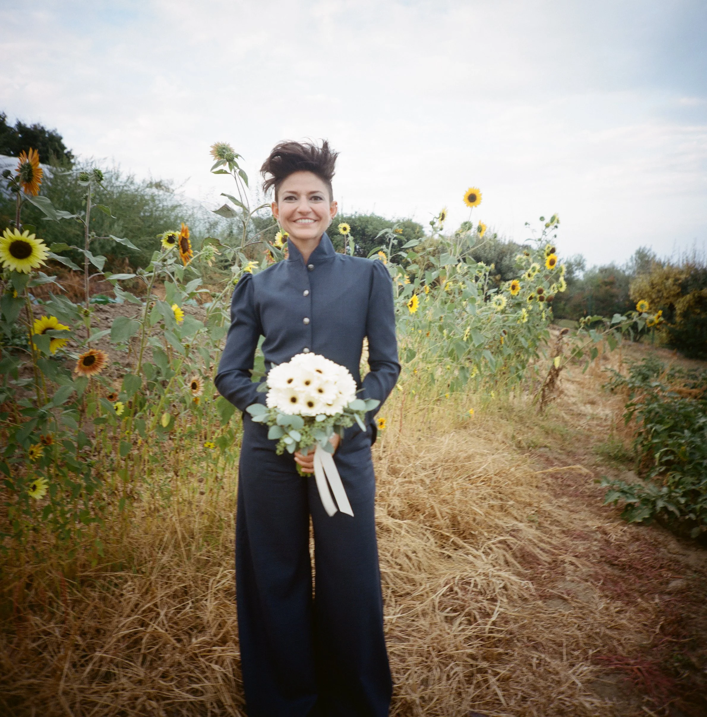 Donna sorridente in un campo di girasoli, indossa un cappotto blu. Lo sfondo mostra il cielo nuvoloso.