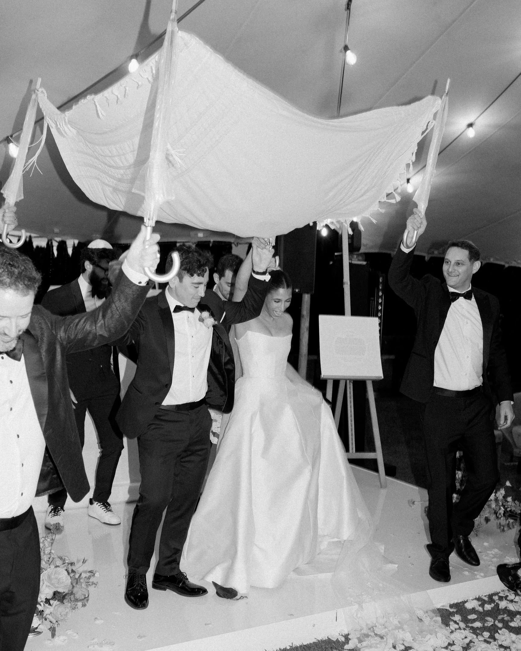 Gruppo di persone che partecipano a una cerimonia di matrimonio, alcuni che tengono un ombrello decorato sopra la coppia di sposi al centro, che indossa abiti eleganti, in un ambiente interno con luci e decorazioni.