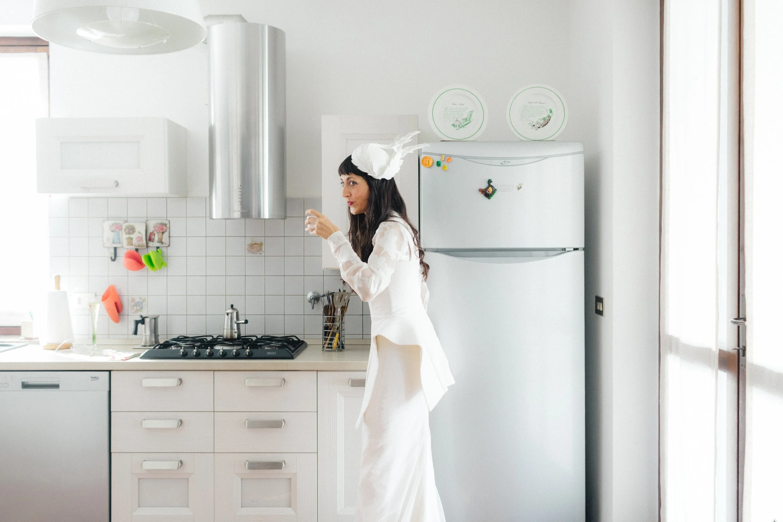 Donna vestita con un costume bianco con Applications di piume indossa un cappello decorato con piume bianche mentre sorseggia un bicchiere in cucina bianca con pensili e frigorifero decorato con magneti colorati e piatti con disegni