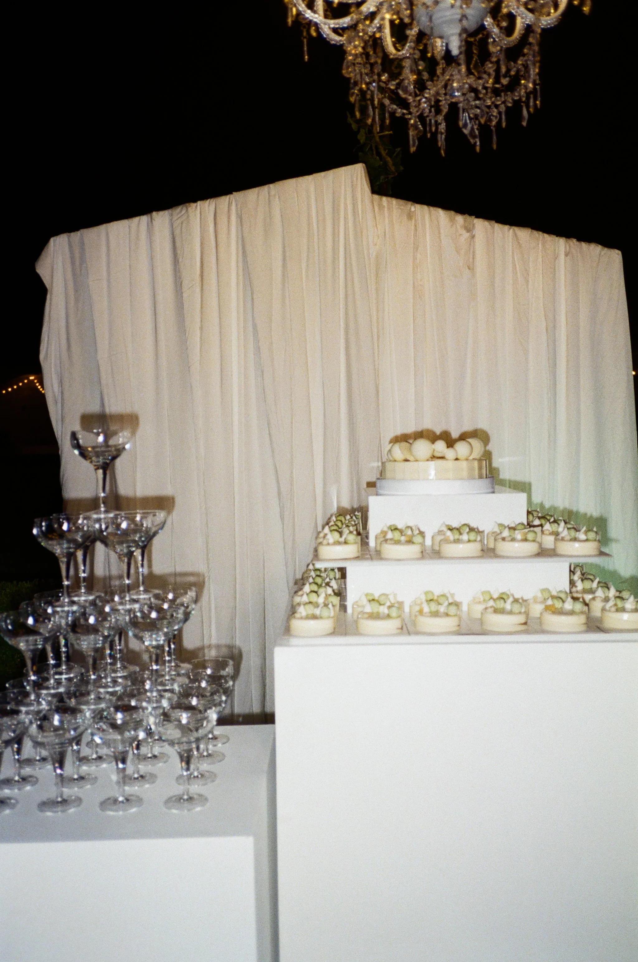 Tavolo con un capitolo di Champagne vuoto e un treft di cristalli. In secondo piano, un tavolino con un grande dolce decorato con panna e mazzi di cioccolato. Sullo sfondo, una tenda bianca e un lampadario dorato.