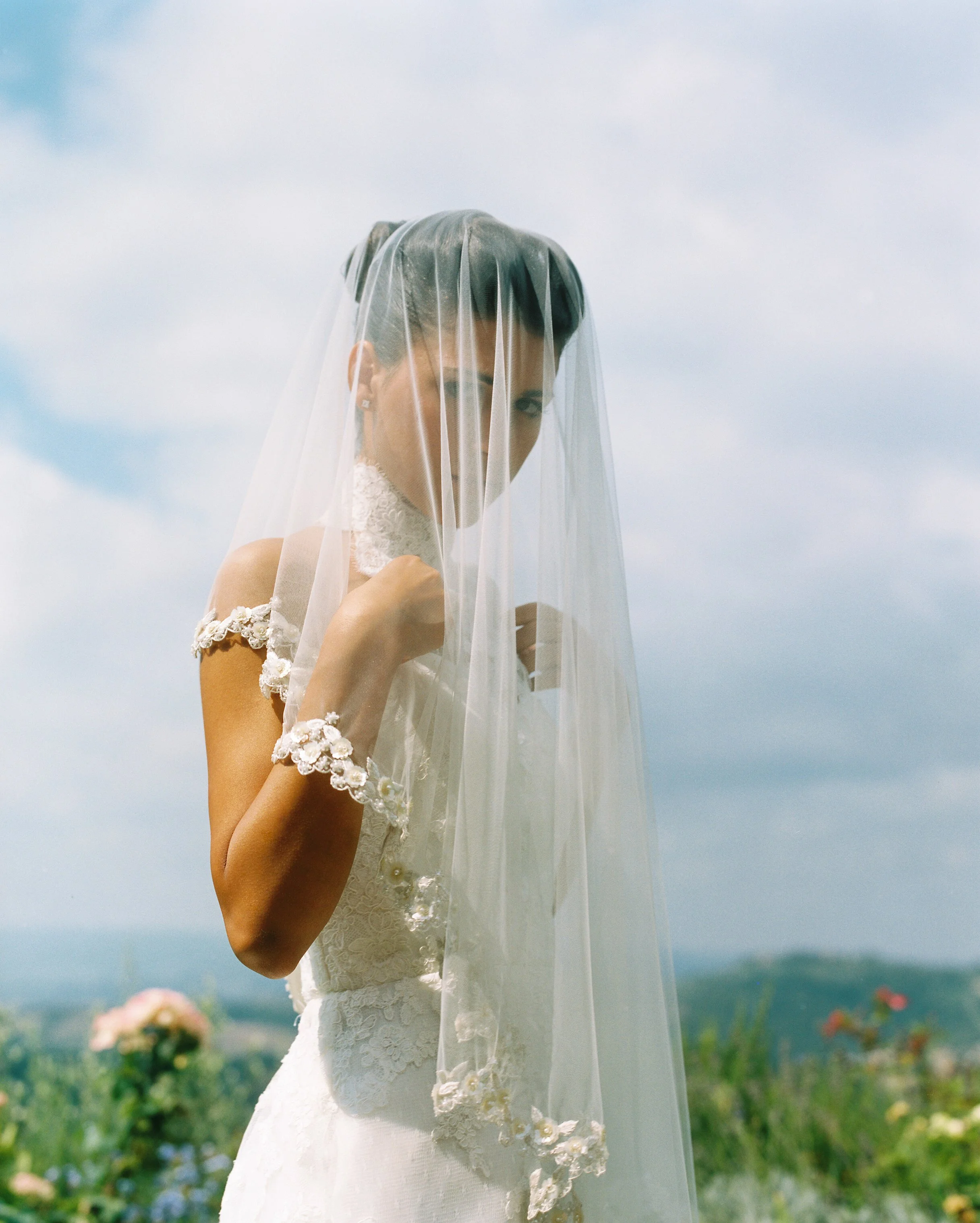 Una donna con un abito di pizzo e un velo bianco si trova in un campo fiorito con il cielo azzurro e nuvoloso.