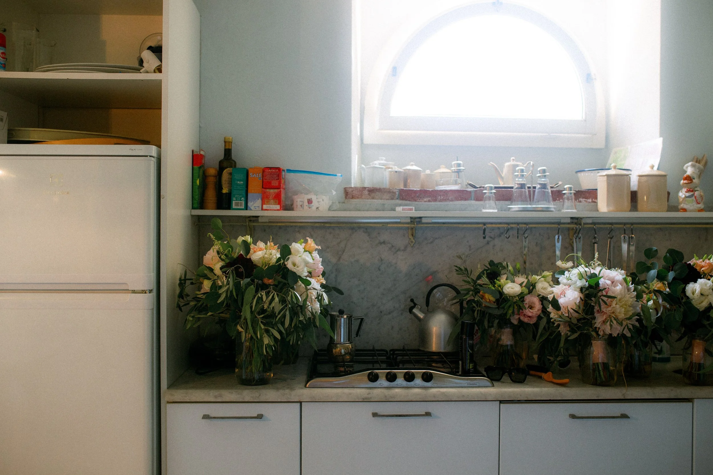 Una cucina con una finestra luminosa, fiori decorativi vicino al piano cottura, e vari utensili e contenitori sul ripiano.