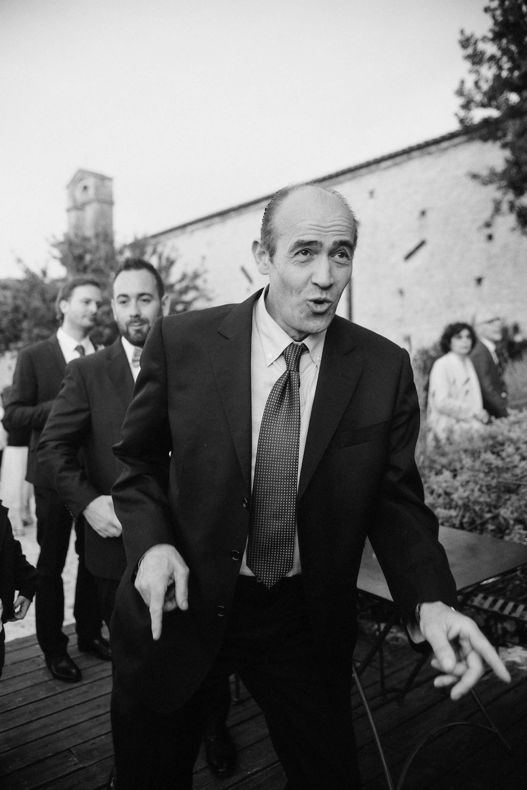 Uomo anziano in abito elegante che suona il pianoforte, circondato da altre persone in abbigliamento formale, in un ambiente all'aperto.