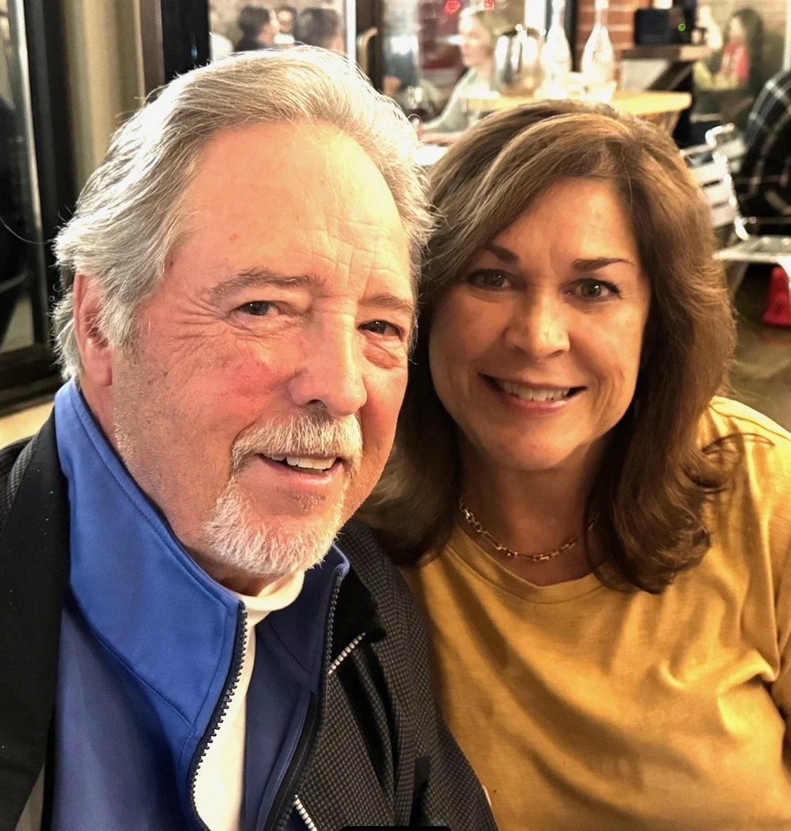 A close-up of an elderly man with gray hair and a beard and a middle-aged woman with shoulder-length brown hair, both smiling at the camera, in a cozy indoor setting.