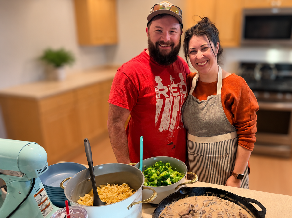 A man and woman smiling in a kitchen with chopped broccoli, pasta, and a skillet of cooked food on the counter.