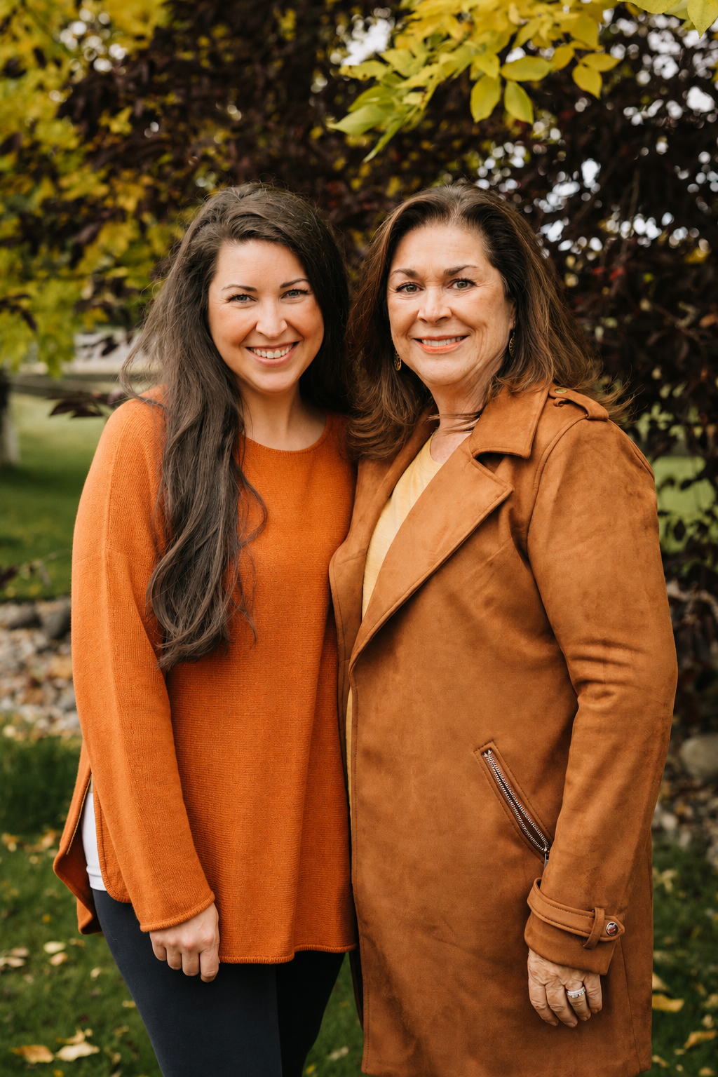 Two women smiling outdoors, standing close together in front of a tree with autumn leaves, wearing orange and brown jackets.