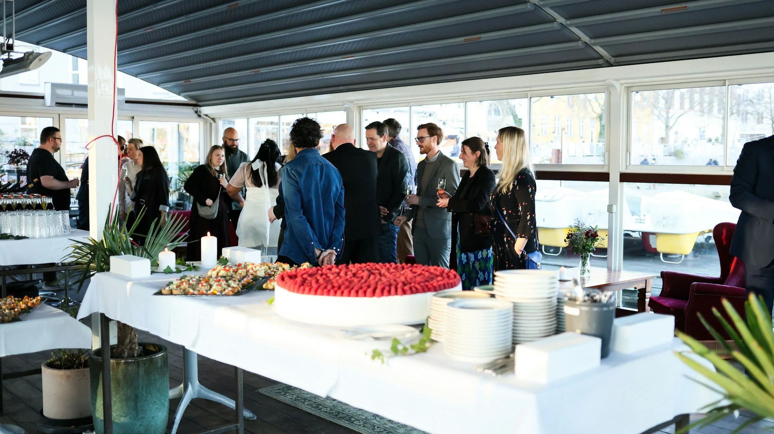 A group of people socializing at a catered corporate event in a bright, modern space with large windows. There is a table with desserts and candles in the foreground, and the attendees are holding drinks and engaging in conversation.