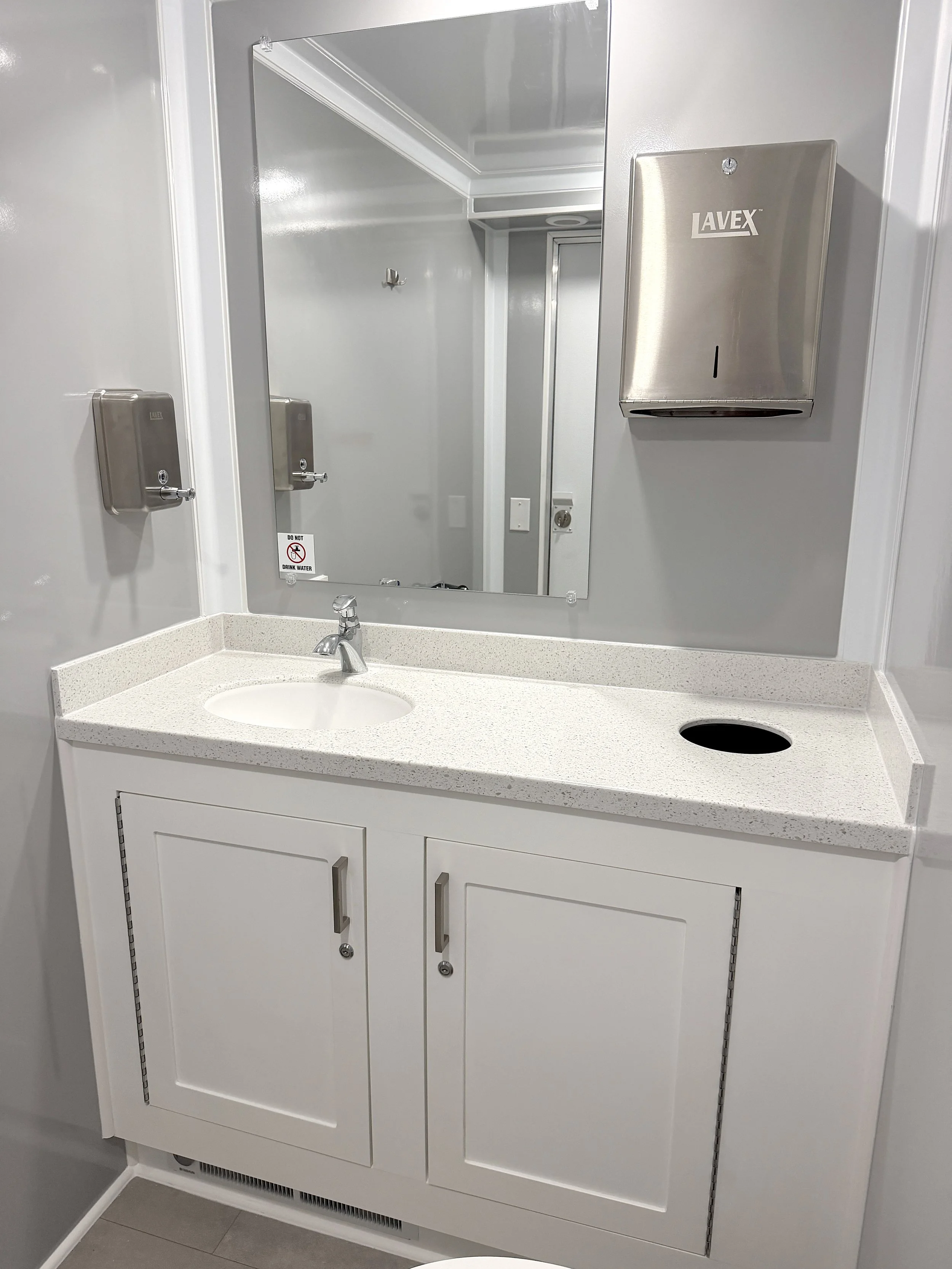 A bathroom with a white vanity cabinet, a mirror, a sink, a soap dispenser, a paper towel dispenser, and a small hole in the counter, with gray tile flooring and gray walls.