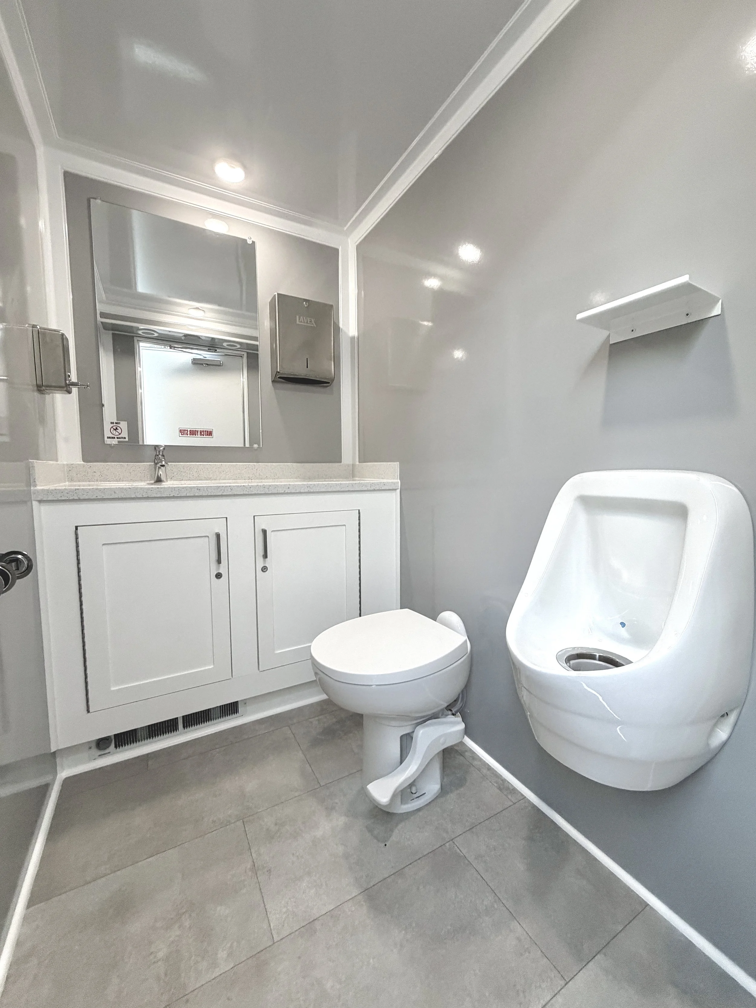 A modern, clean bathroom with gray tiled floor, a white toilet, a white sink with a cabinet below, a mirror above the sink, a metal paper towel dispenser, a wall-mounted soap or sanitizer dispenser, and a wall-mounted white shelf. There is a sign on the mirror indicating no smoking or water use, and the bathroom appears to be in a mobile or compact space.