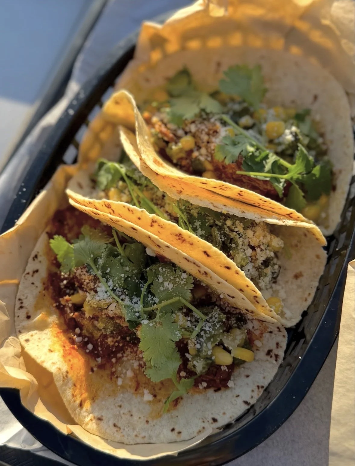 Three tacos in a black basket, garnished with cilantro, with grilled tortillas and various fillings.