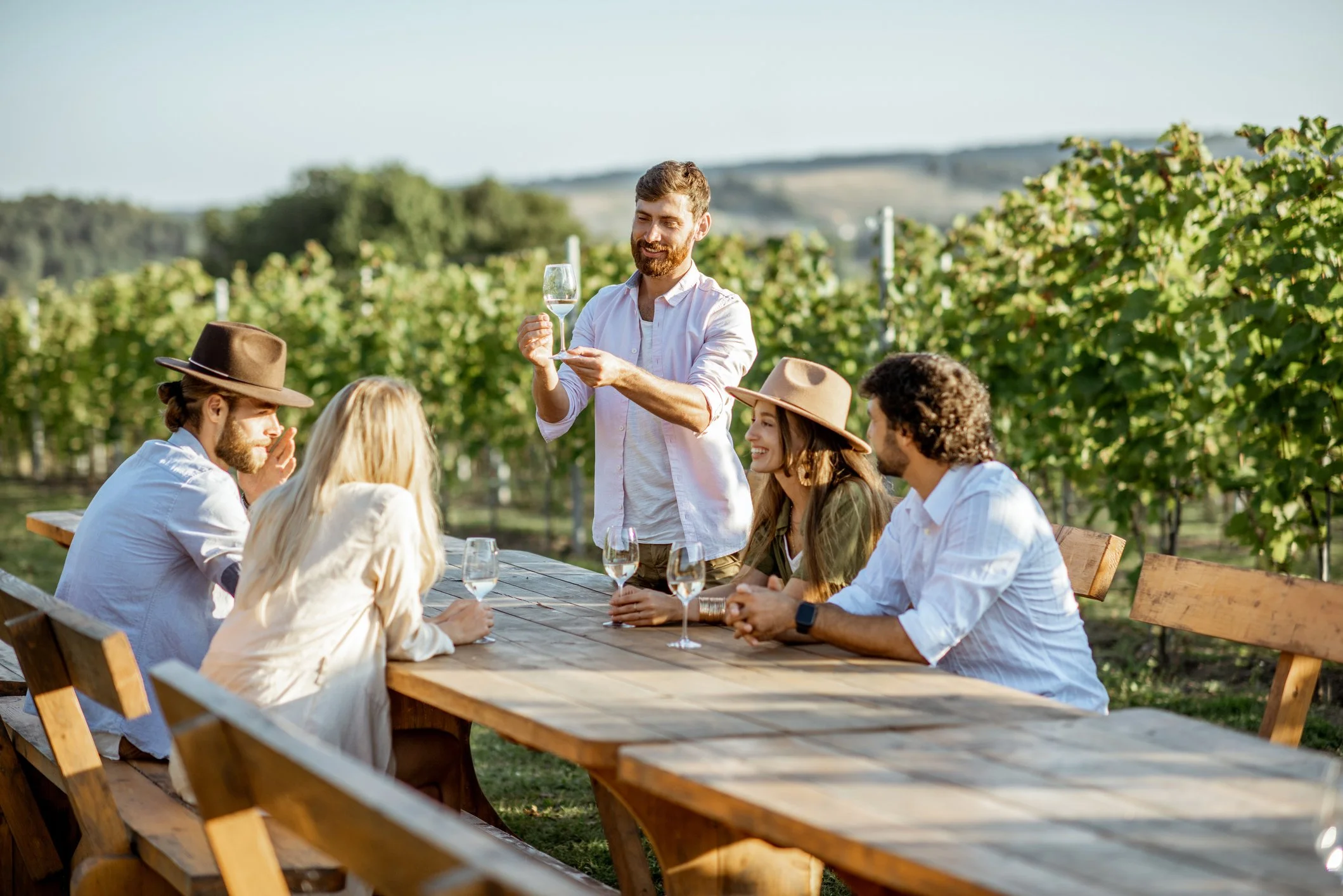 A group of people outdoors at a vineyard, with one person standing holding a wine glass while others sit at a wooden table, surrounded by lush green grapevines under a clear sky.