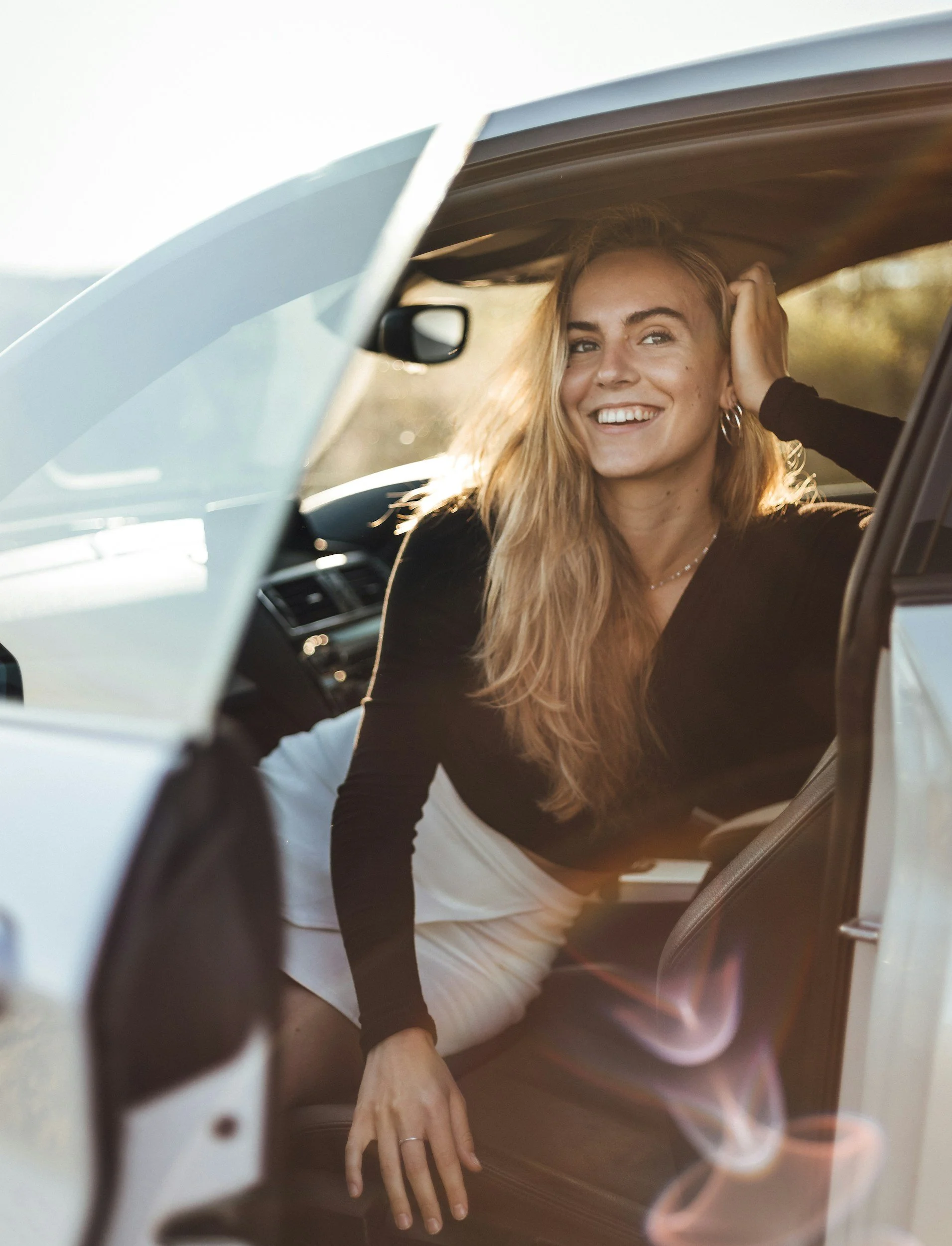 A woman with blonde hair smiling inside a car, leaning toward the open door with her hand resting on the car's interior.