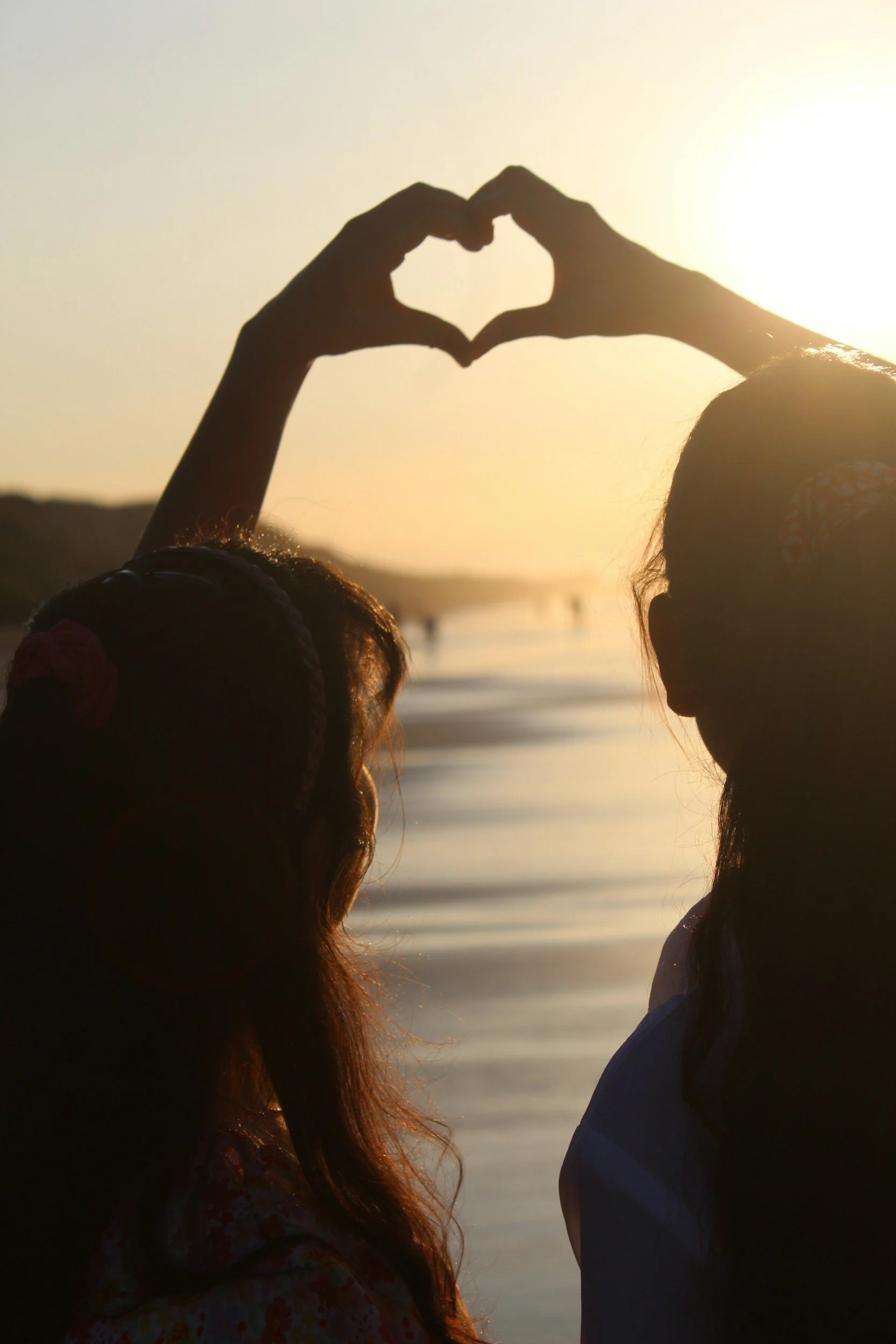 Silhouette of two children at the beach during sunset, forming a heart with their hands.