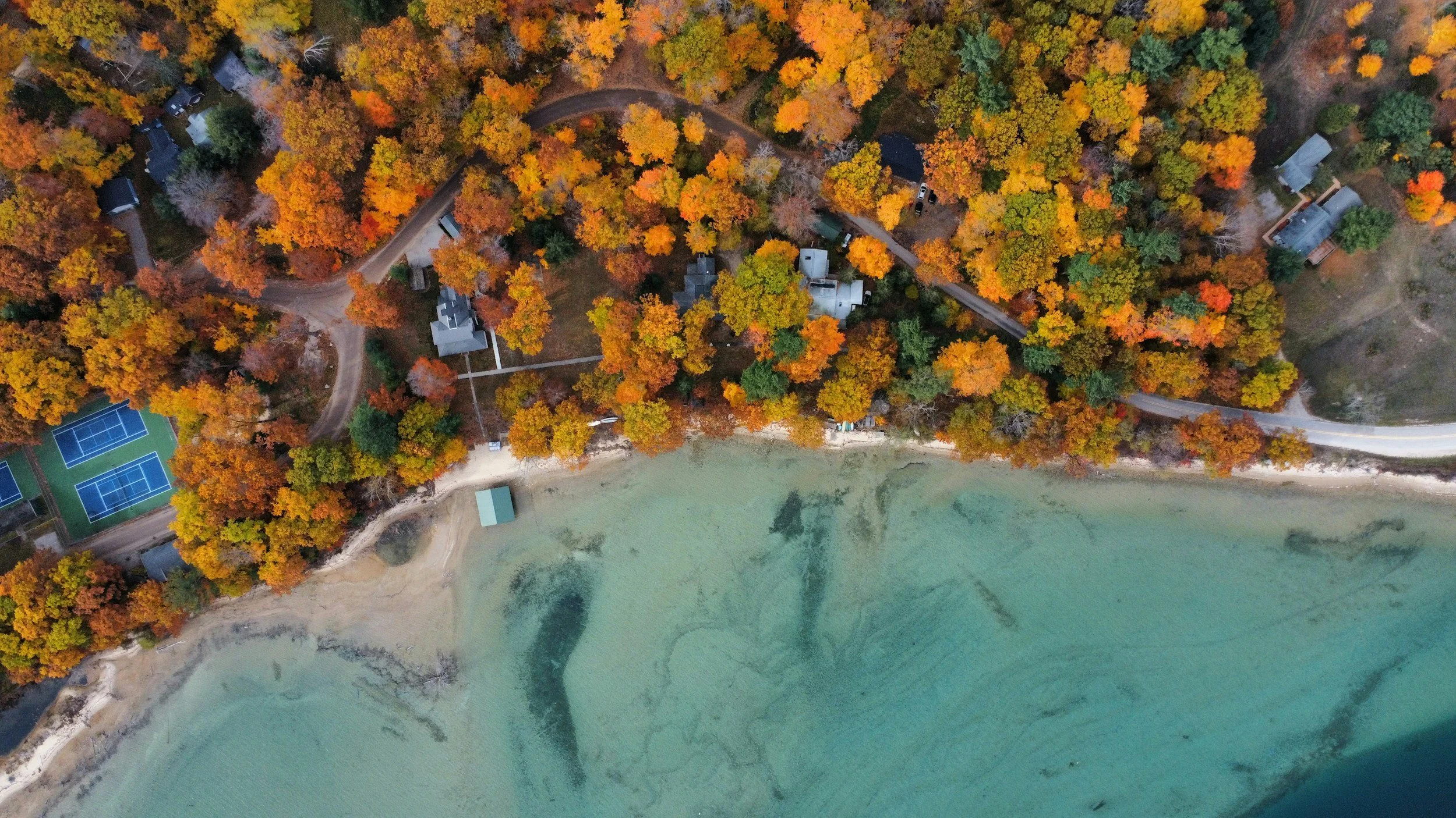An aerial view of a lakeside area with a forest showing fall foliage, winding roads through the trees, houses, and tennis courts.