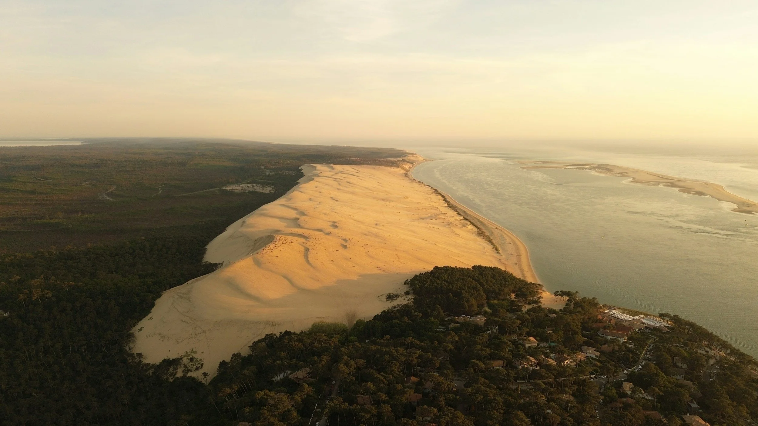 Aerial view of a sandy coastal barrier island with greenery, sand dunes, and nearby residential area, leading to the ocean.