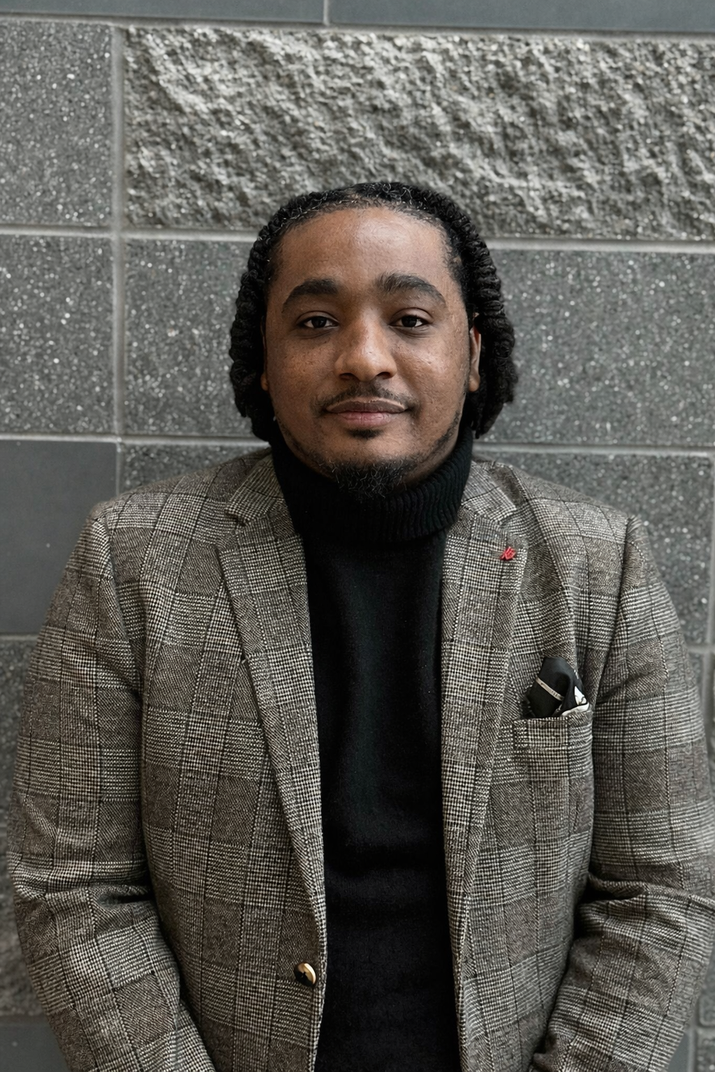 A man with dark skin, black hair styled in twists, wearing a brown plaid blazer over a black turtleneck, seated against a gray concrete block wall.