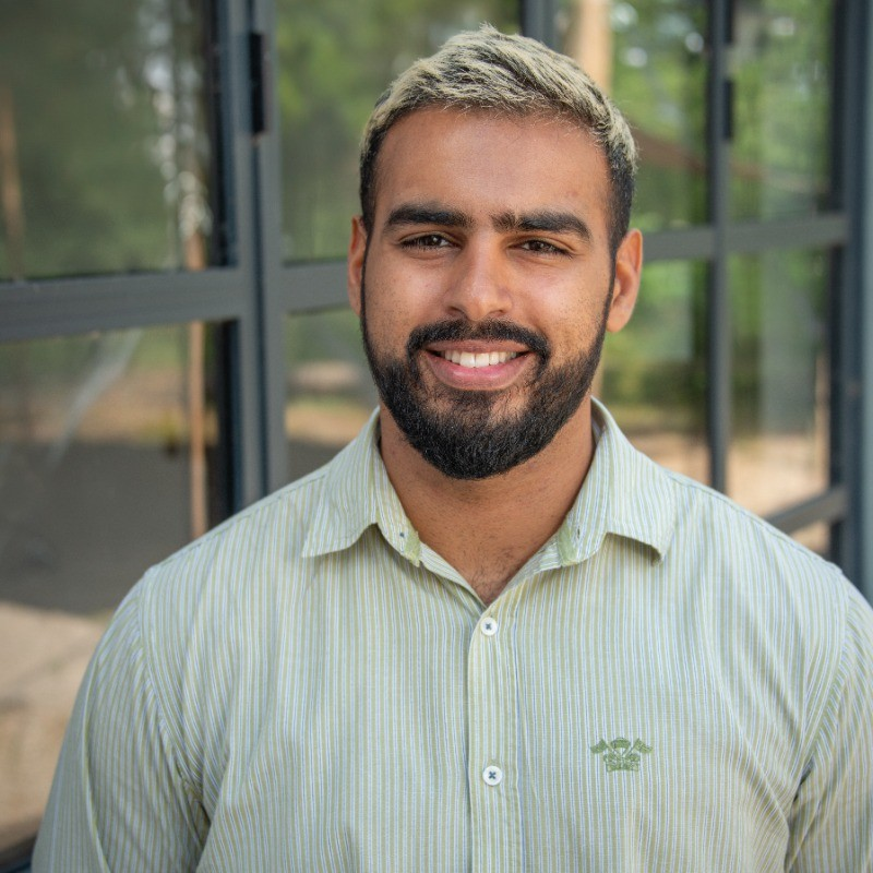 A smiling man with a beard and short hair, wearing a light green striped button-up shirt, standing outdoors near a glass building with green trees in the background.