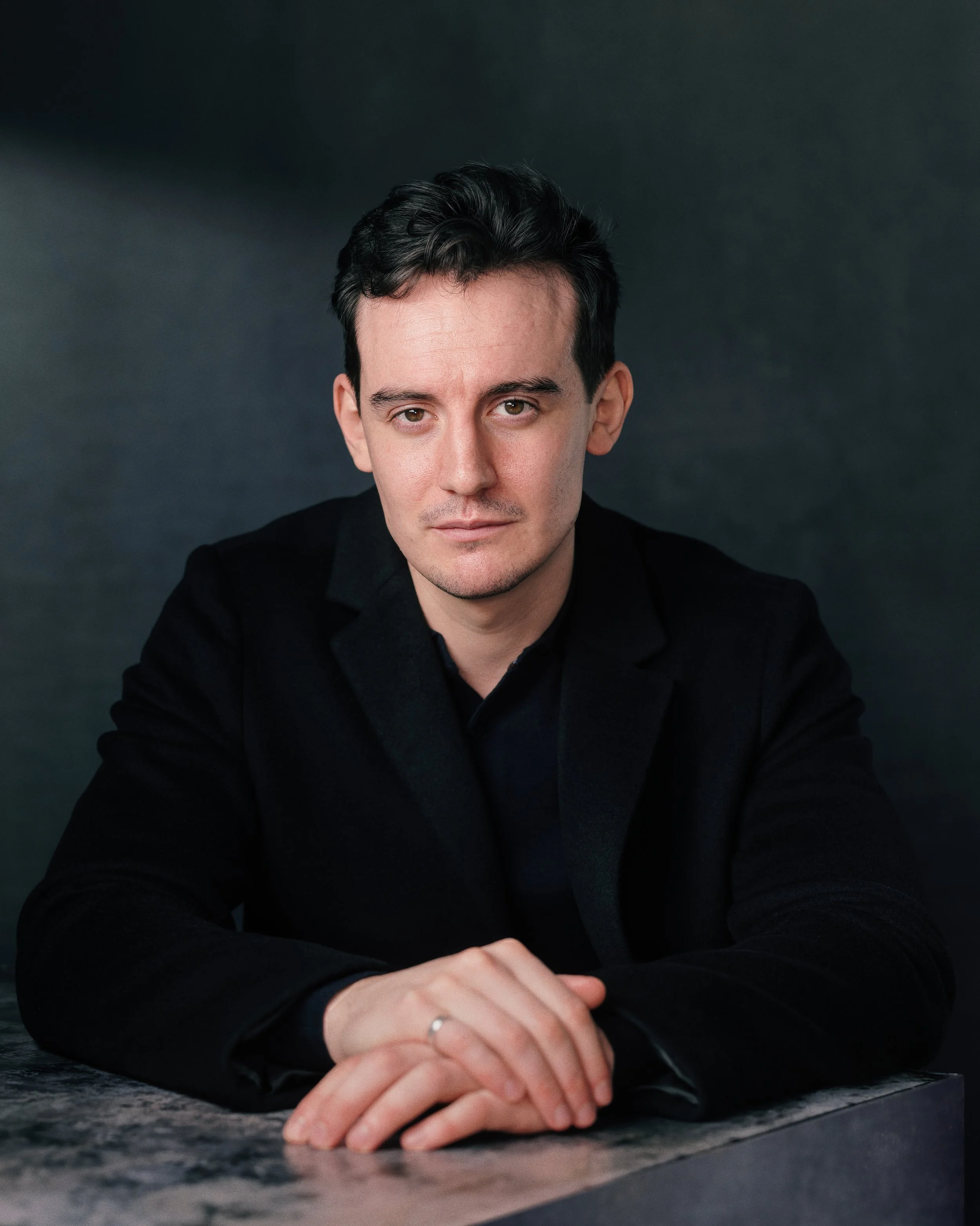 A man with dark hair and light skin, wearing a black shirt, leans on a gray stone surface, with his hands clasped together. The background is dark and blurred.