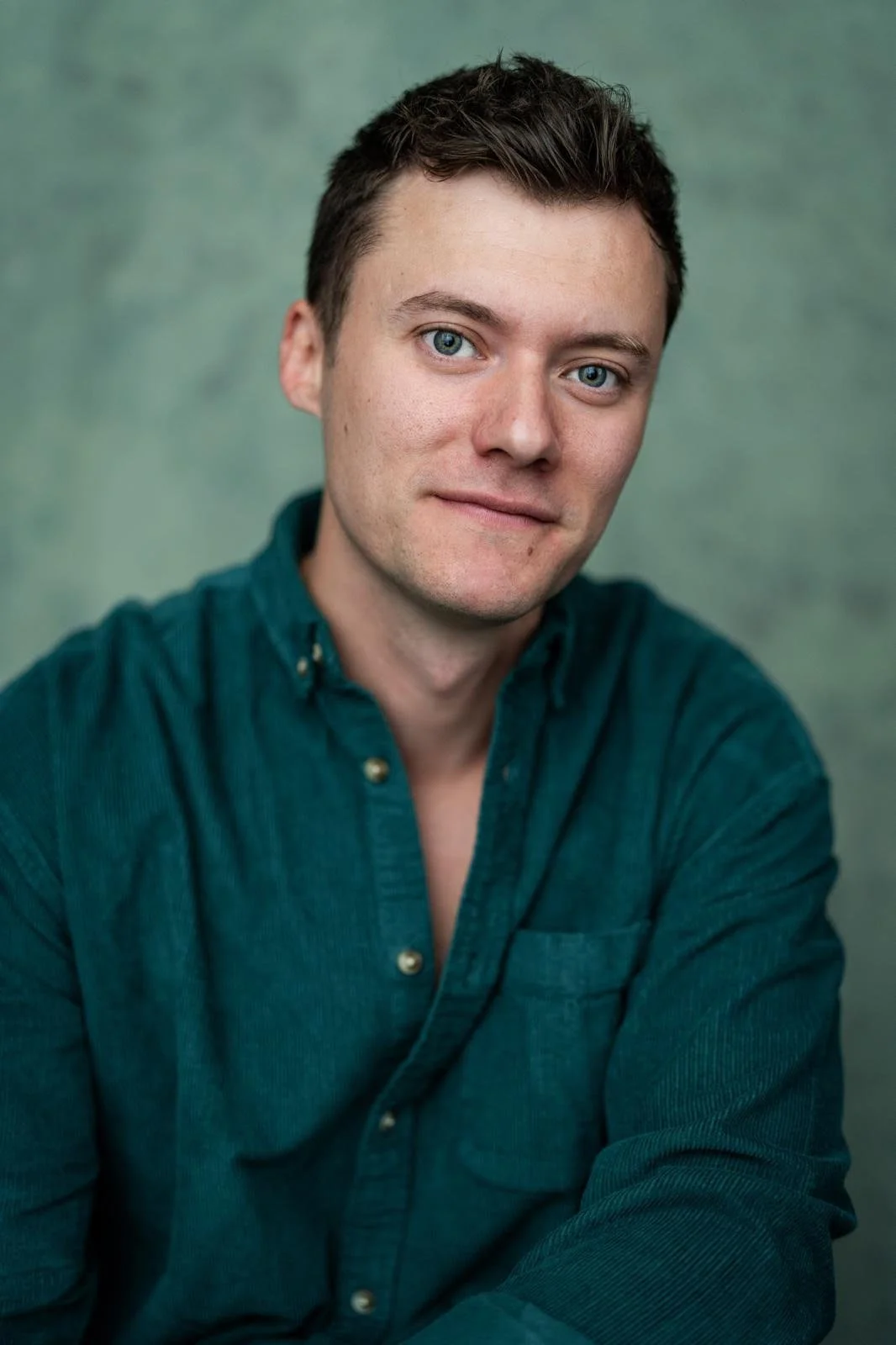 Portrait of a young man with short dark hair and blue eyes, wearing a teal button-up shirt, sitting against a green backdrop.